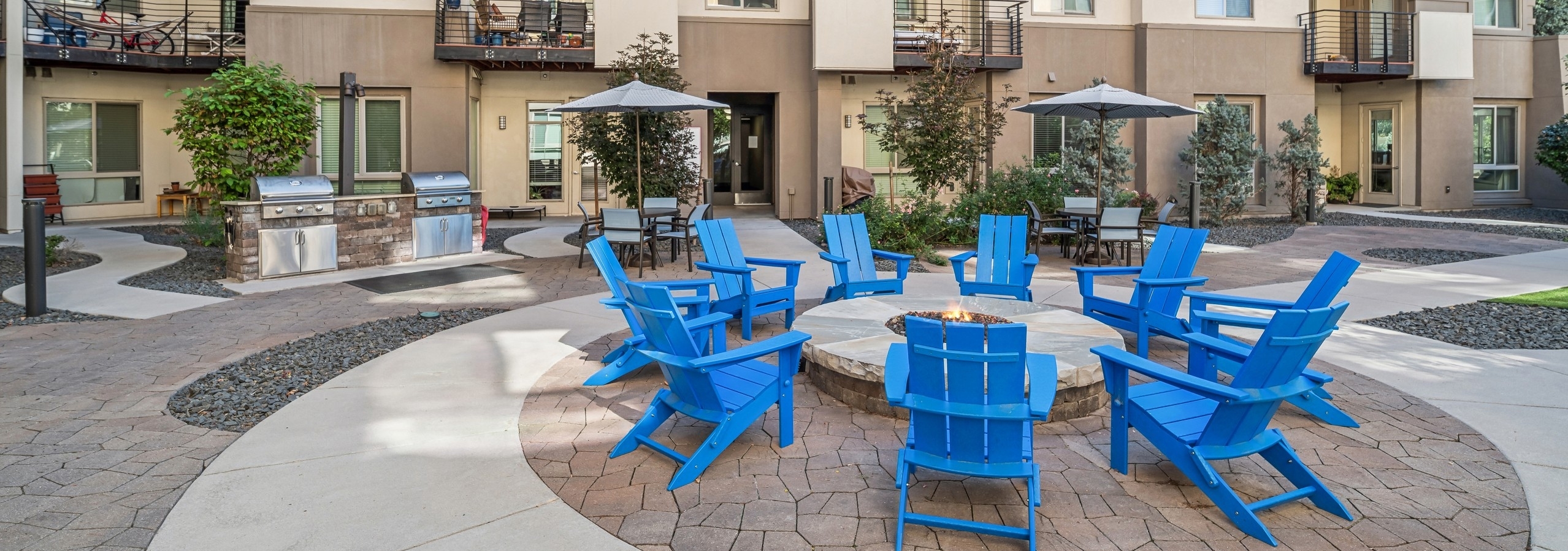 Courtyard fire pit at AMLI Cherry Creek apartments surrounded by blue Adirondack chairs and with grill, tables and chairs in the rear