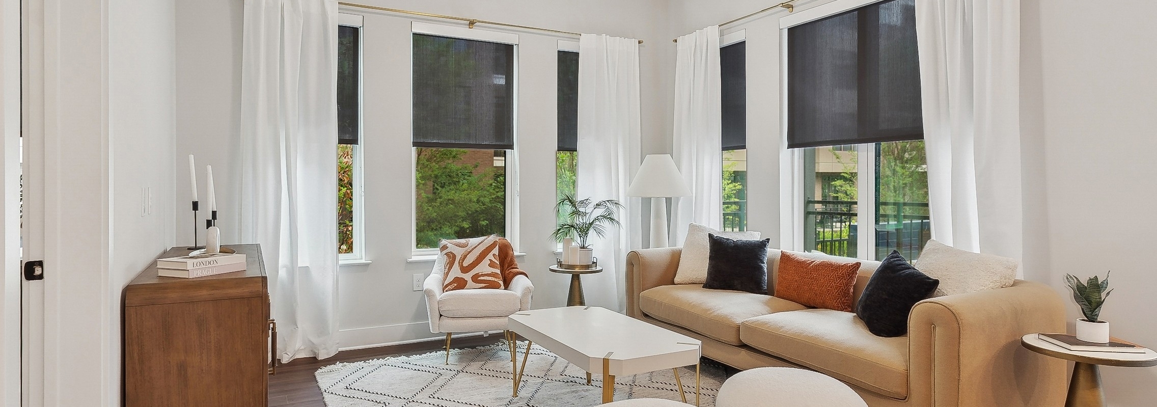 Living room at AMLI Flatiron featuring a tan sofa and white armchair around a white coffee table and white curtains and black roller shades