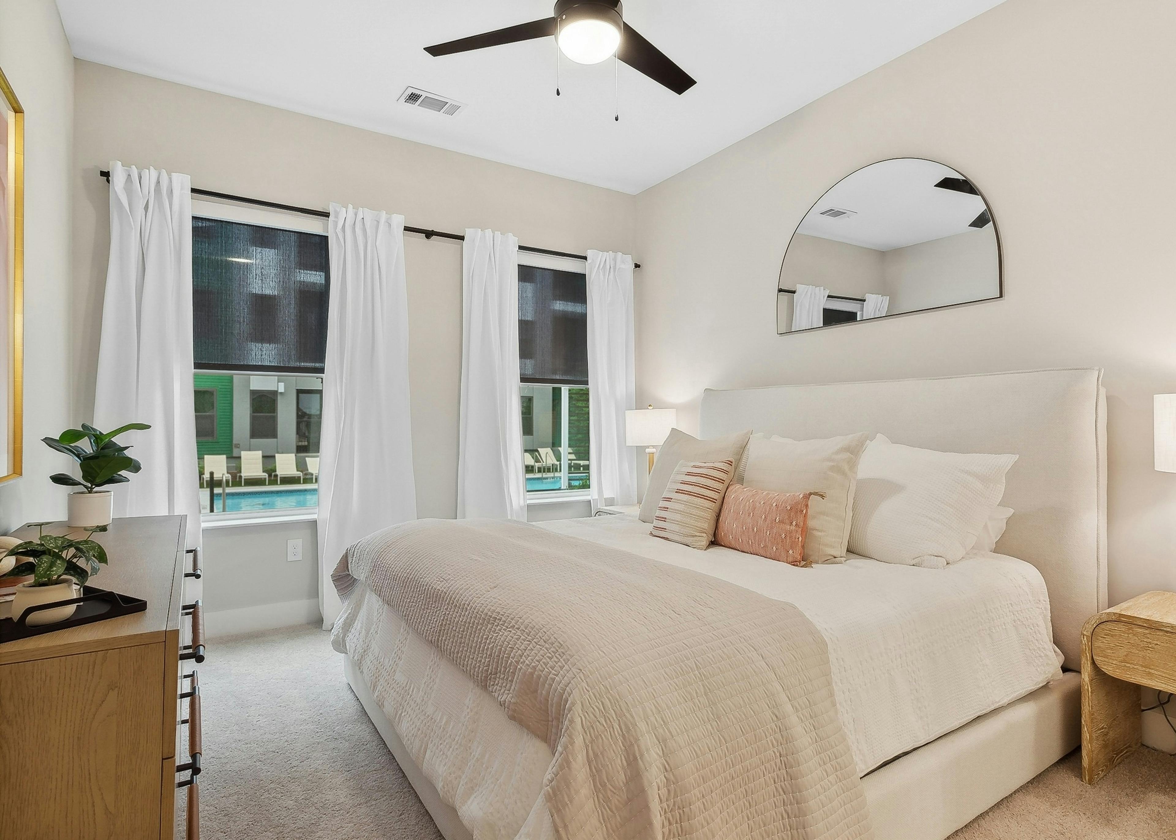 A bedroom at AMLI Flatiron with neutral tones featuring an upholstered bed with tan bedding and several pillows and two large windows