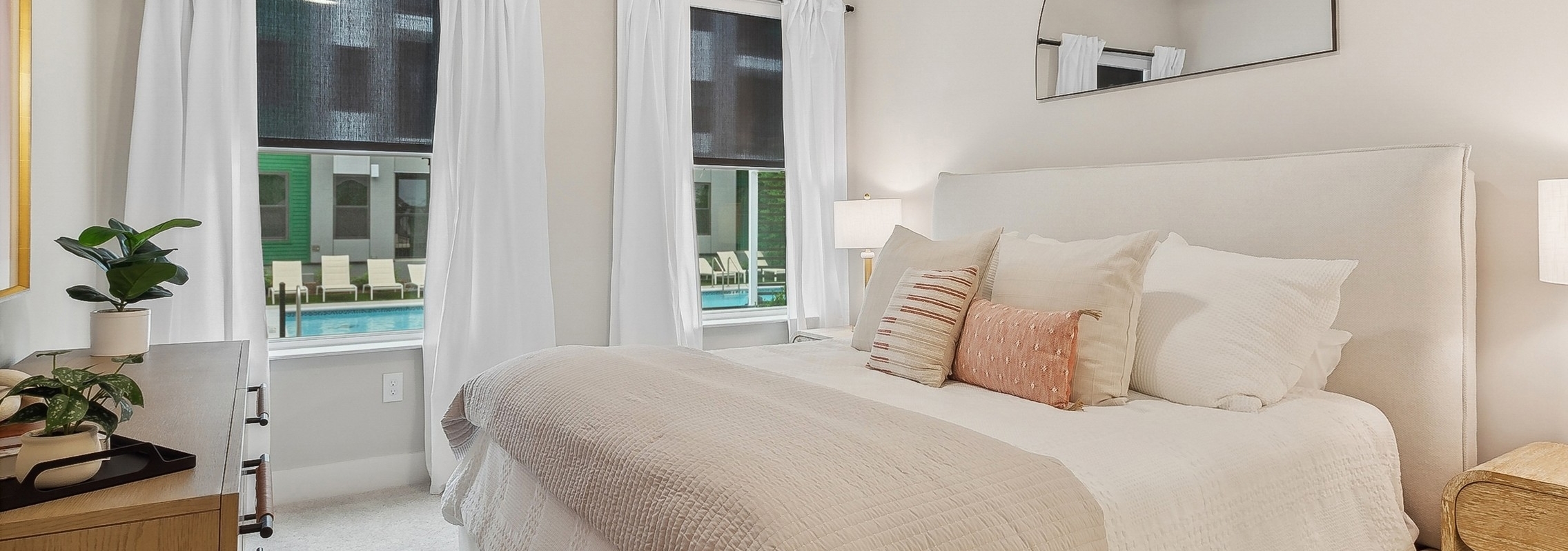 A bedroom at AMLI Flatiron with neutral tones featuring an upholstered bed with tan bedding and several pillows and two large windows