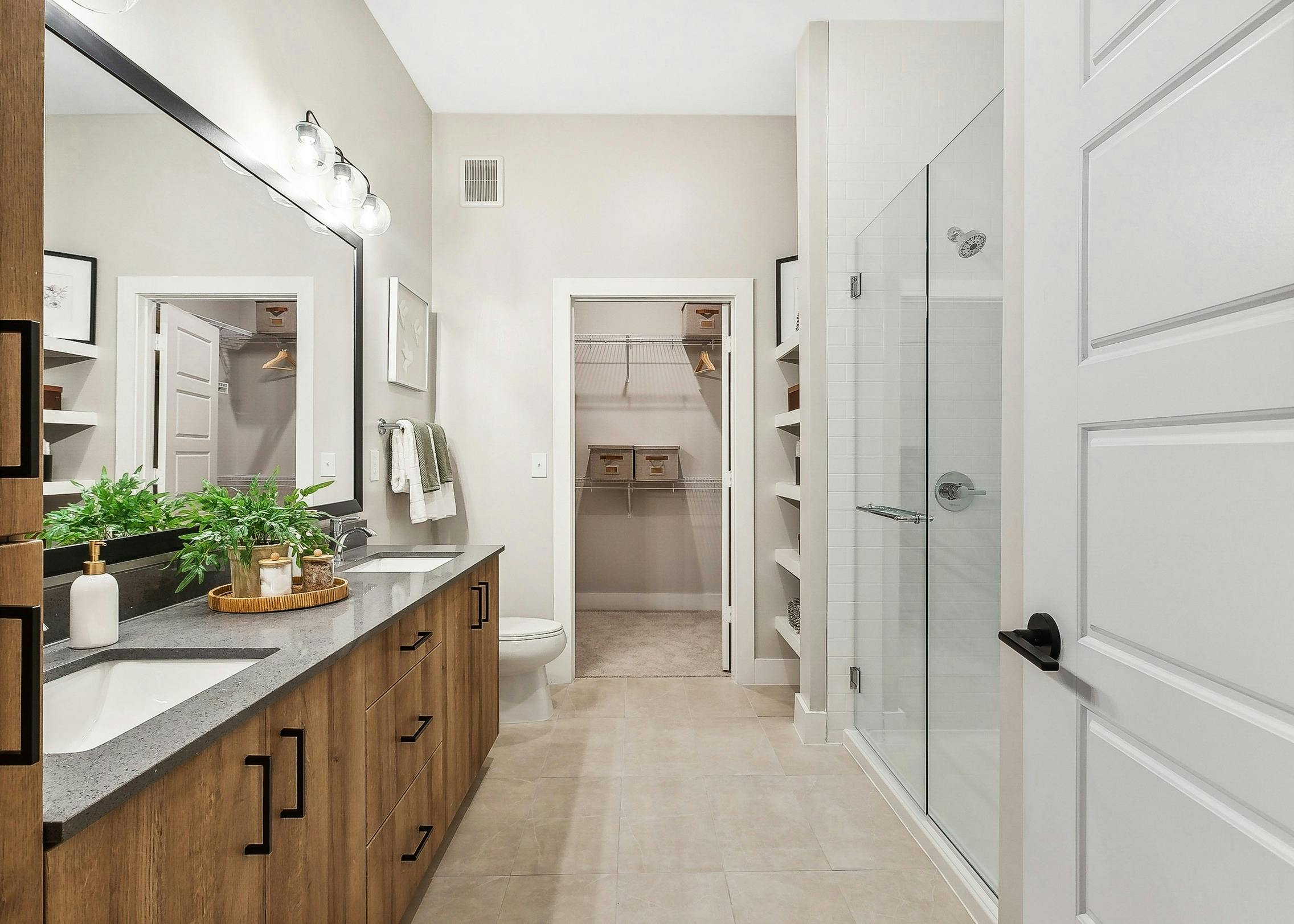 Modern bathroom at AMLI Flatiron featuring a double wooden vanity with a large mirror above and glass shower with view into walk-in closet