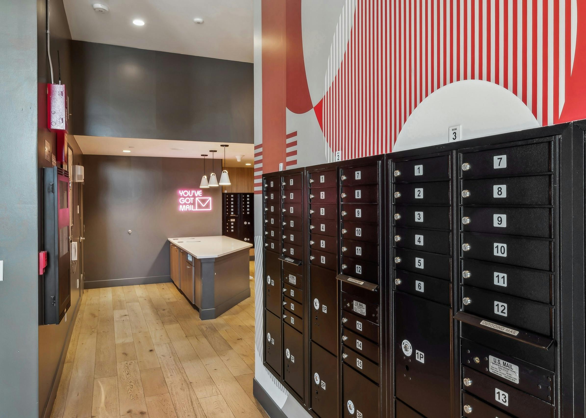 Black numbered mailboxes at AMLI Broadway Park and a neon sign reading "You've Got Mail" above small countertop space in back of mailroom