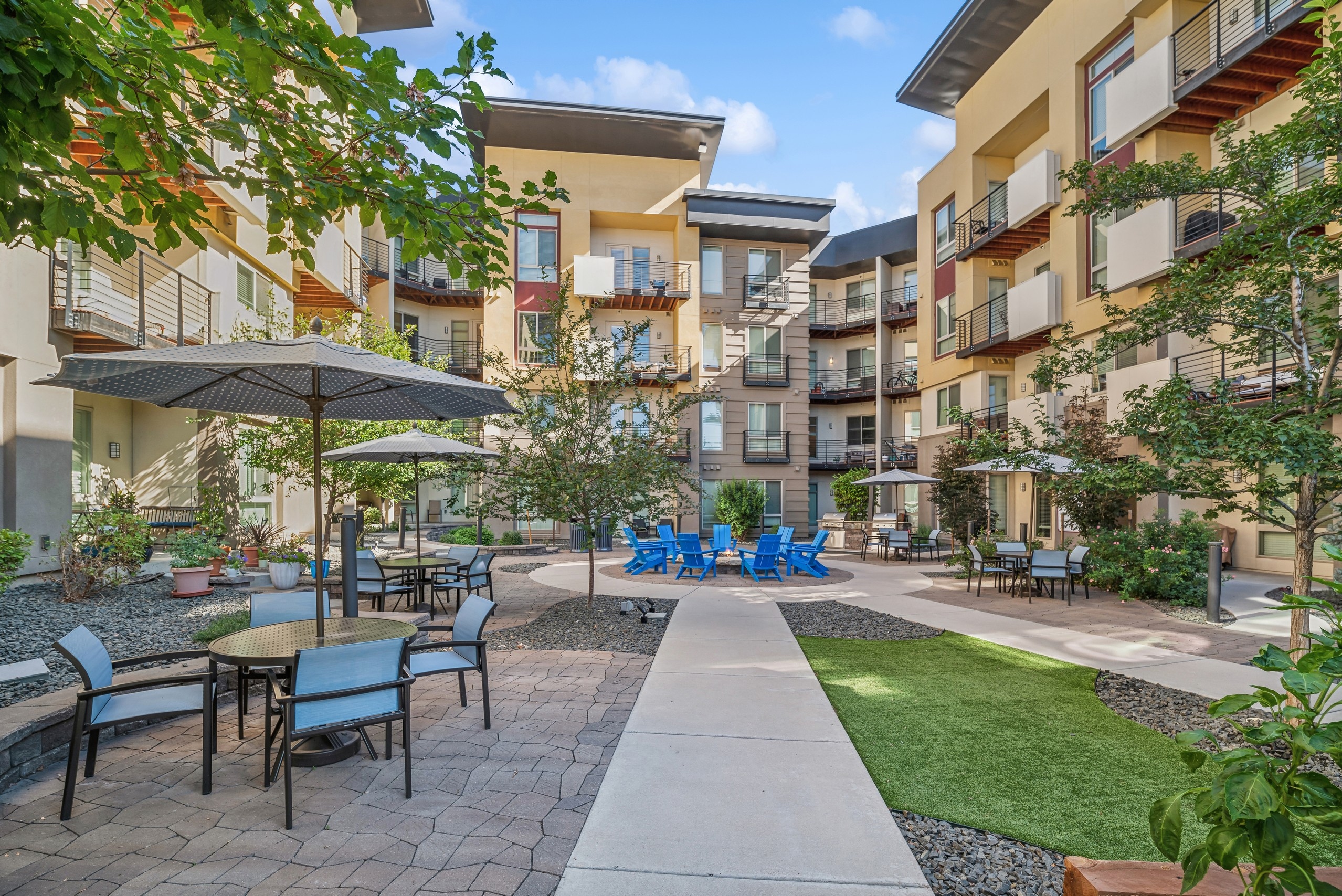 AMLI Cherry Creek courtyard with many trees and umbrella tables and chairs surrounding a fire pit with blue Adirondack chairs