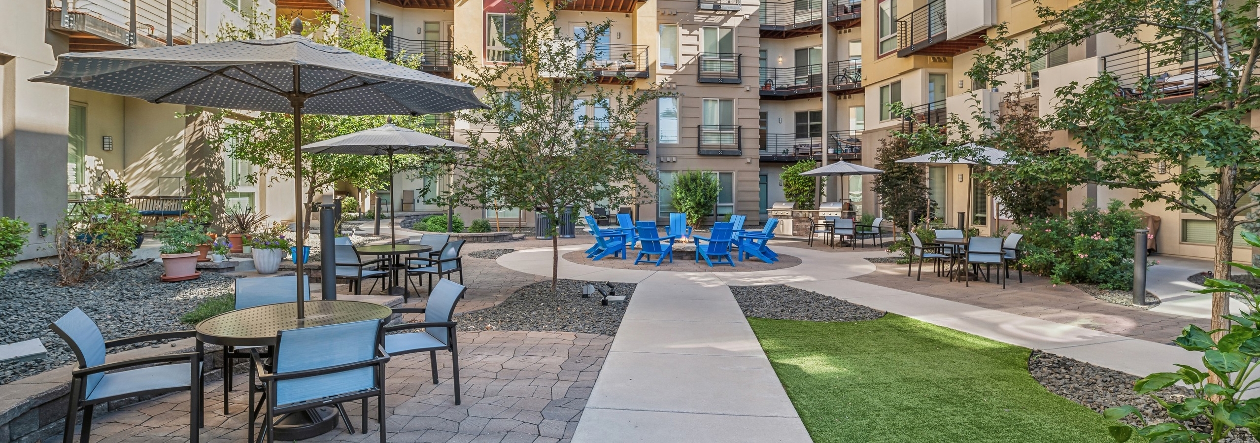 AMLI Cherry Creek courtyard with many trees and umbrella tables and chairs surrounding a fire pit with blue Adirondack chairs