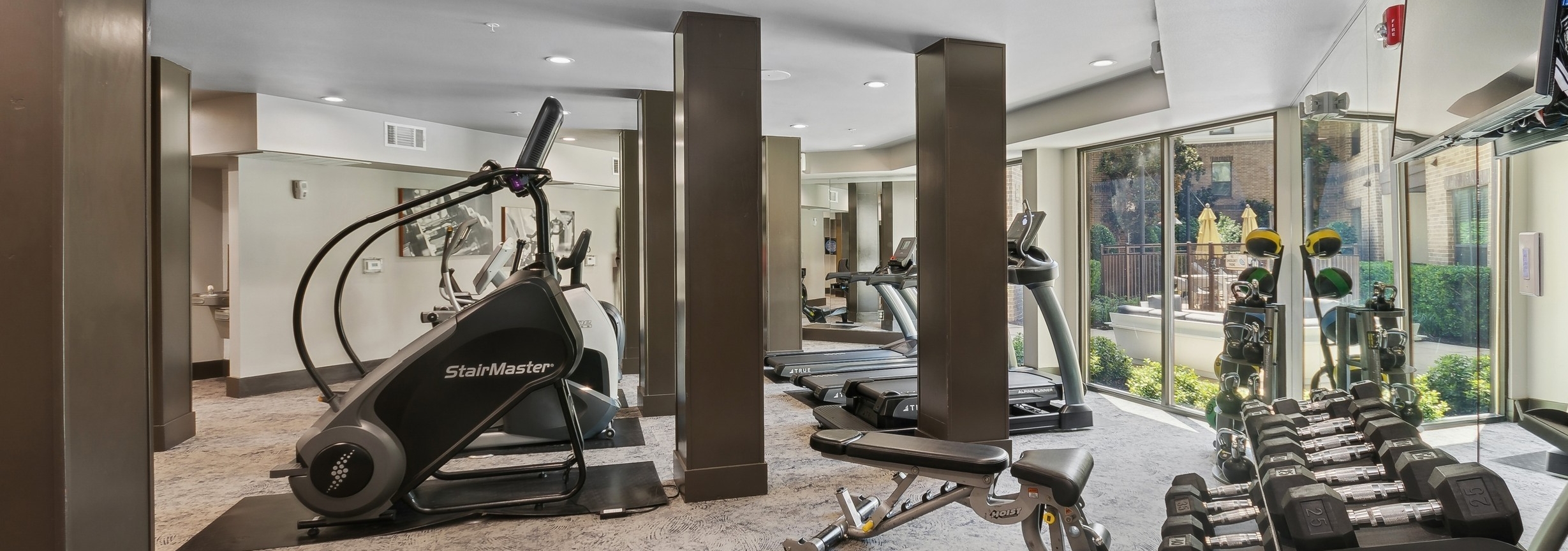 Interior of fitness center at AMLI at the Ballpark apartments with fitness equipment, rack of weights, dark wood pillars and large windows