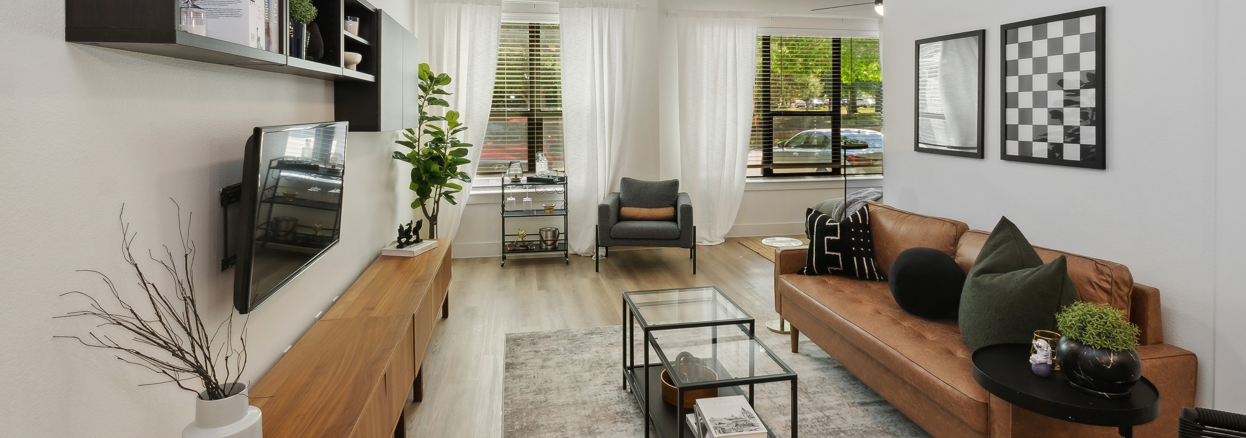 Living room at AMLI at the Ballpark with a brown leather sofa, glass coffee table, a wall-mounted TV with shelves above and large windows