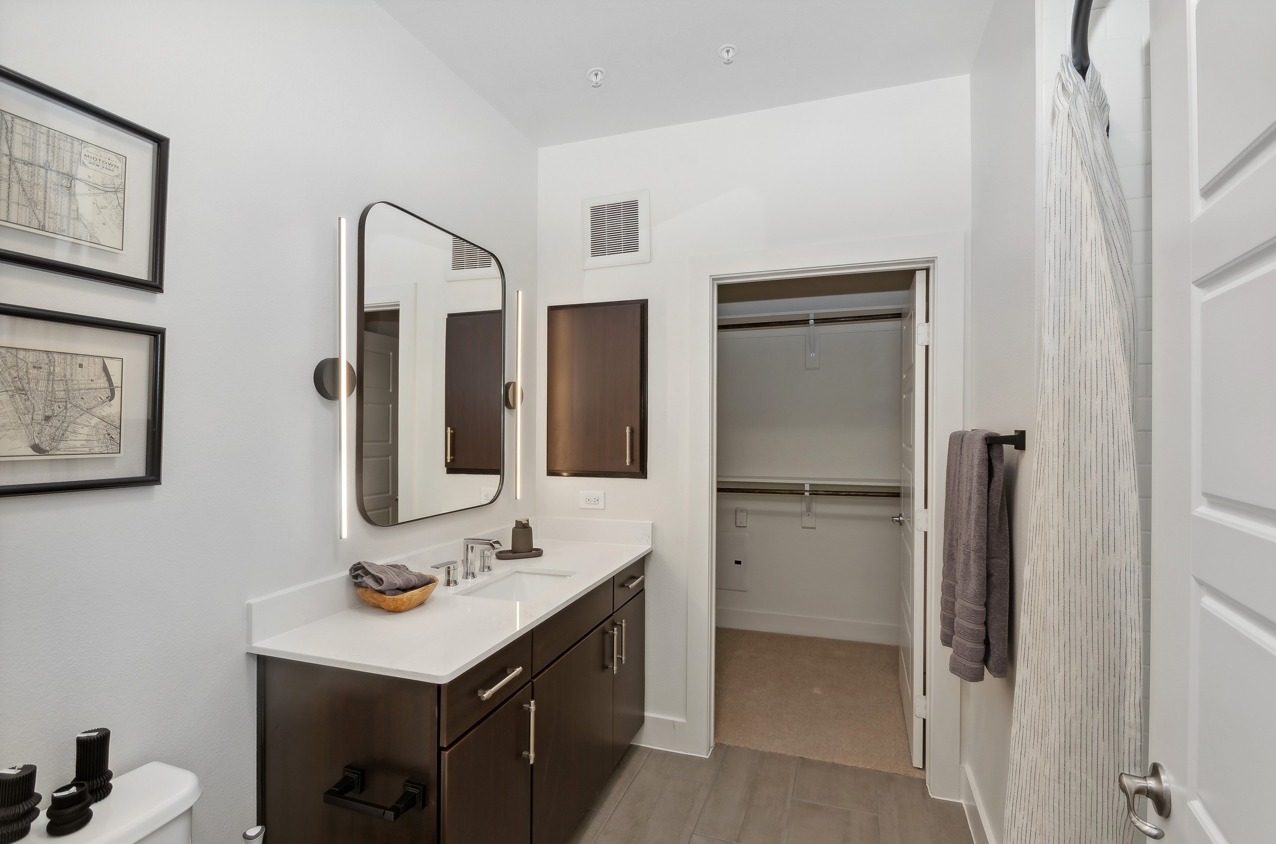 Bathroom at AMLI at the Ballpark with dark wood vanity with white countertop and a large mirror above and a view into walk-in closet