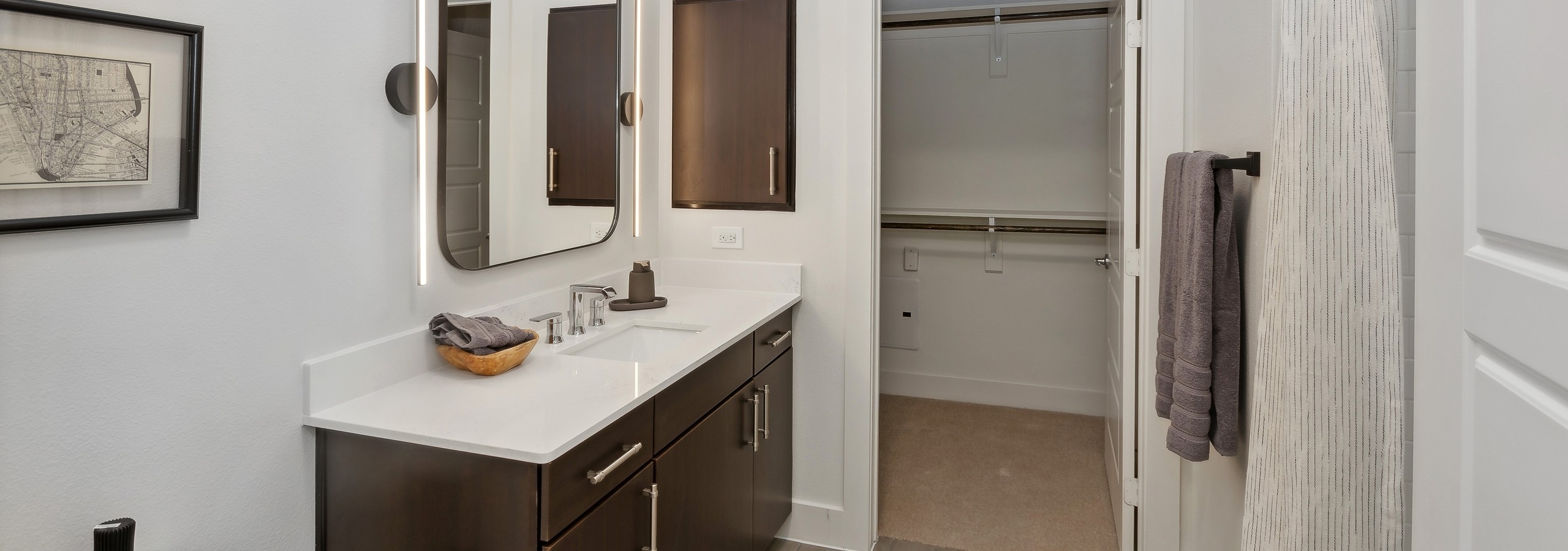 Bathroom at AMLI at the Ballpark with dark wood vanity with white countertop and a large mirror above and a view into walk-in closet