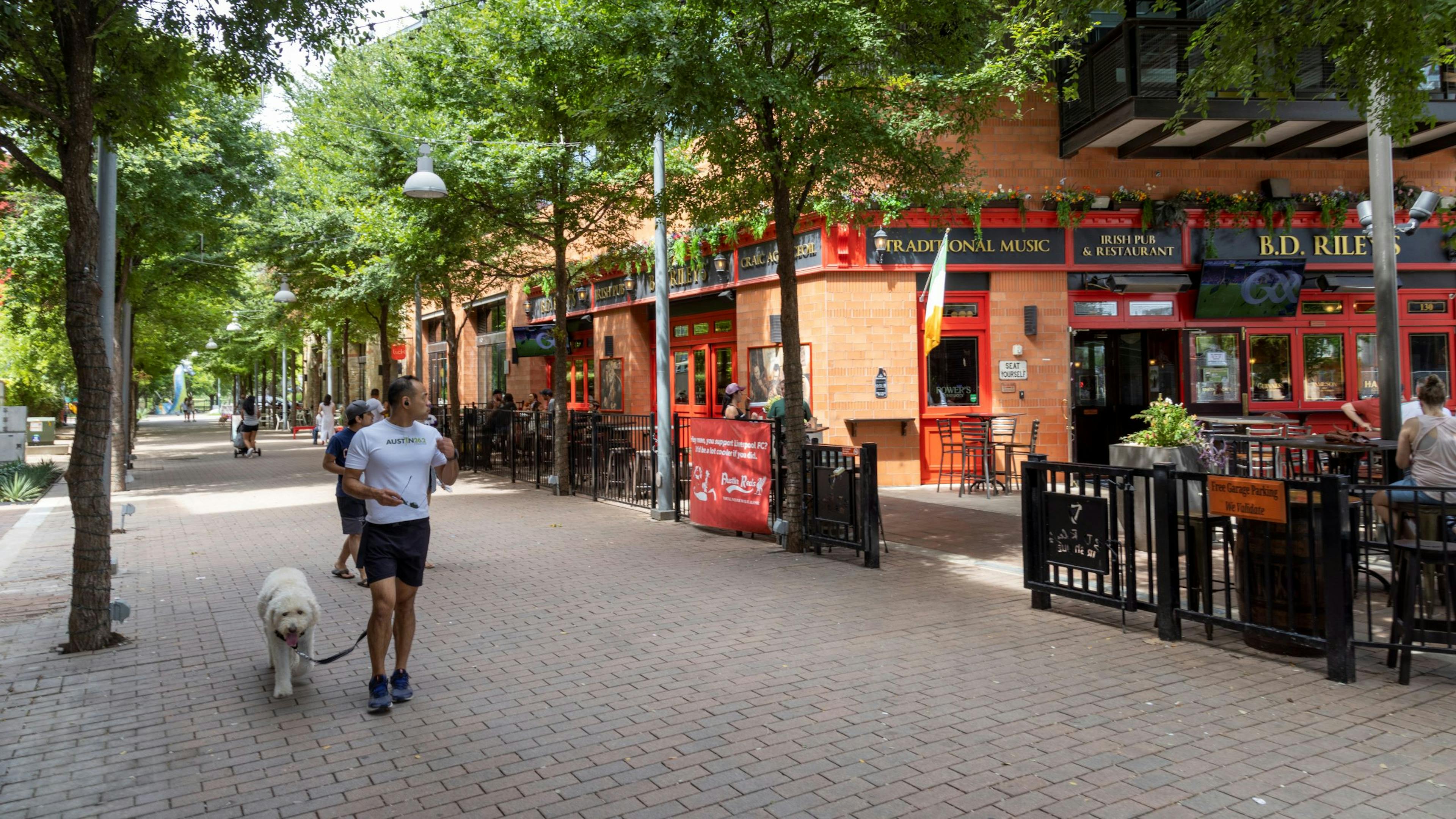 Mueller brick sidewalk lined with trees and man walking his dog near an Irish pub with outdoor and indoor dining