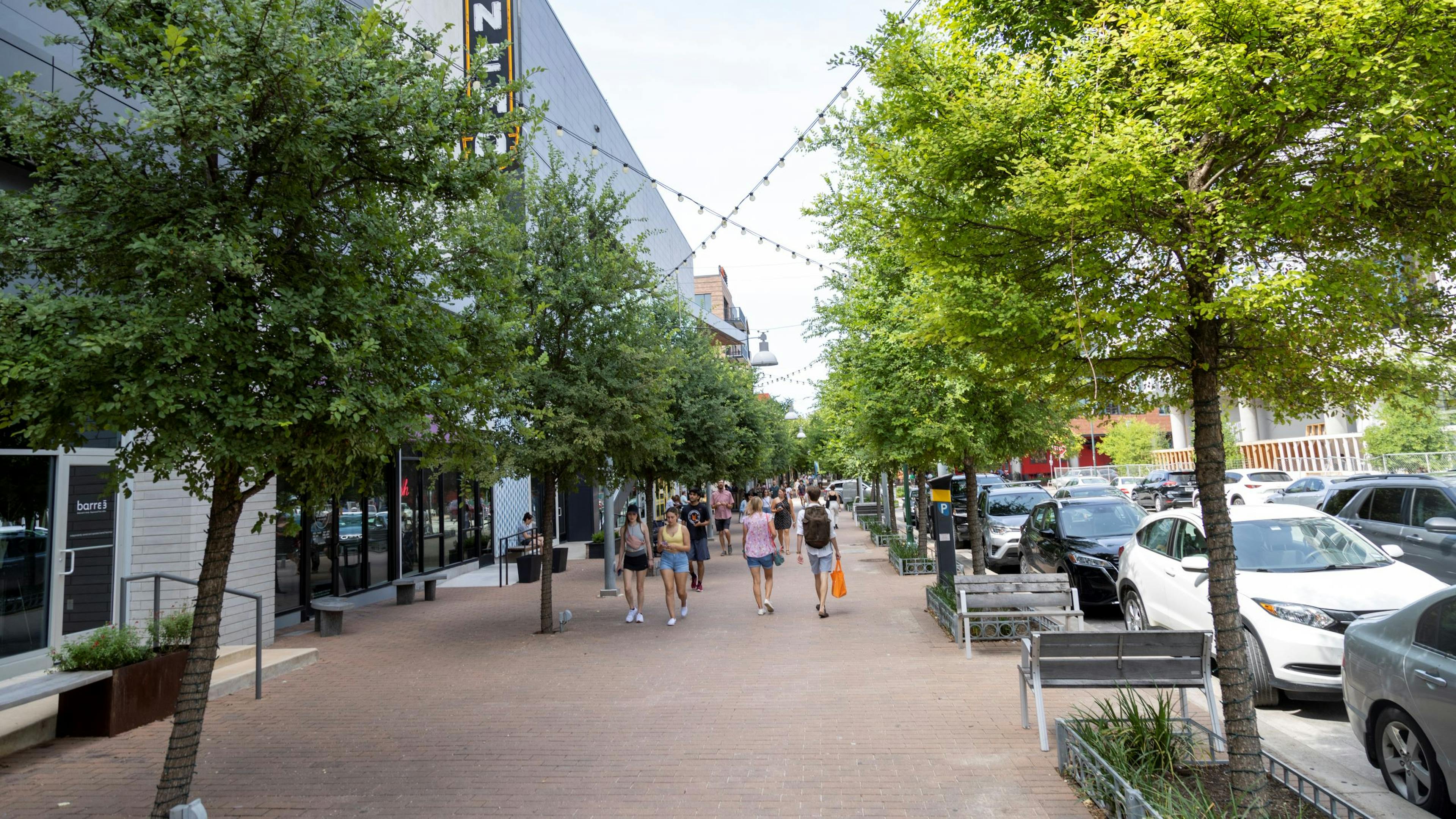 Mueller brick sidewalk lined with trees and  people walking and street with cars parked on both sides