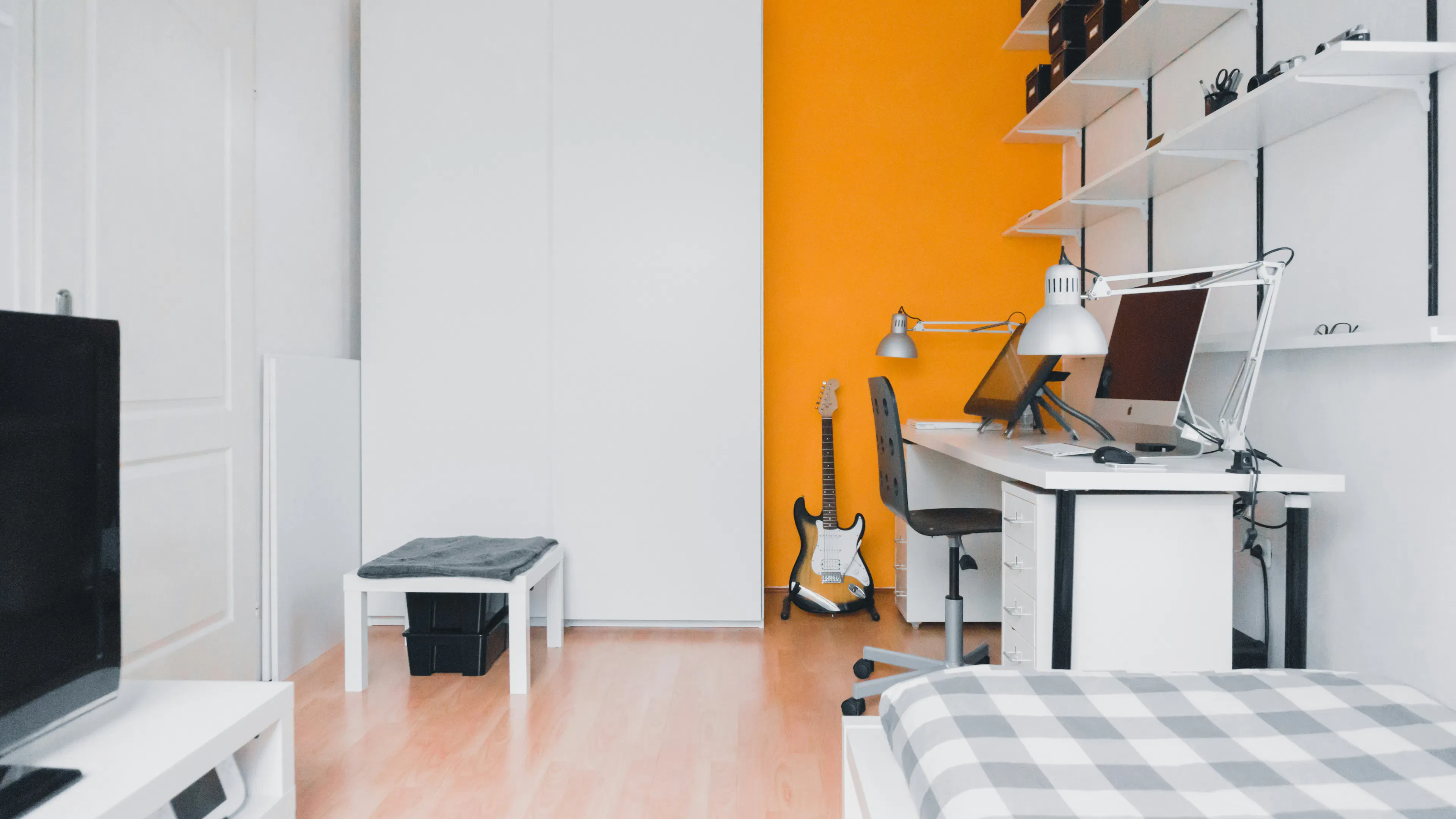 A white desk with a desktop and a guitar next to it in a clean white room with an orange accent wall