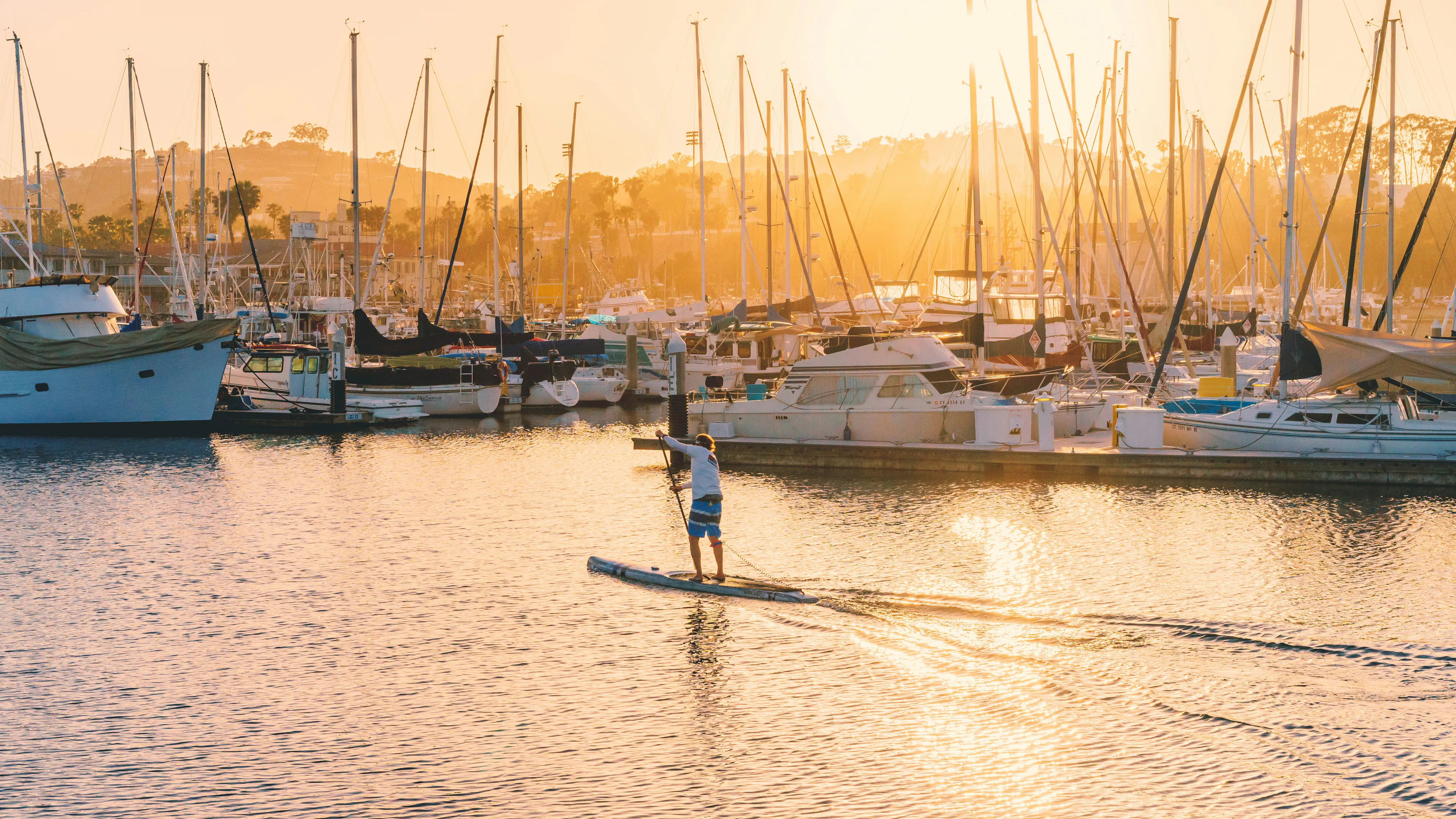 A stand up paddle boarder gliding past some boats in the sunset