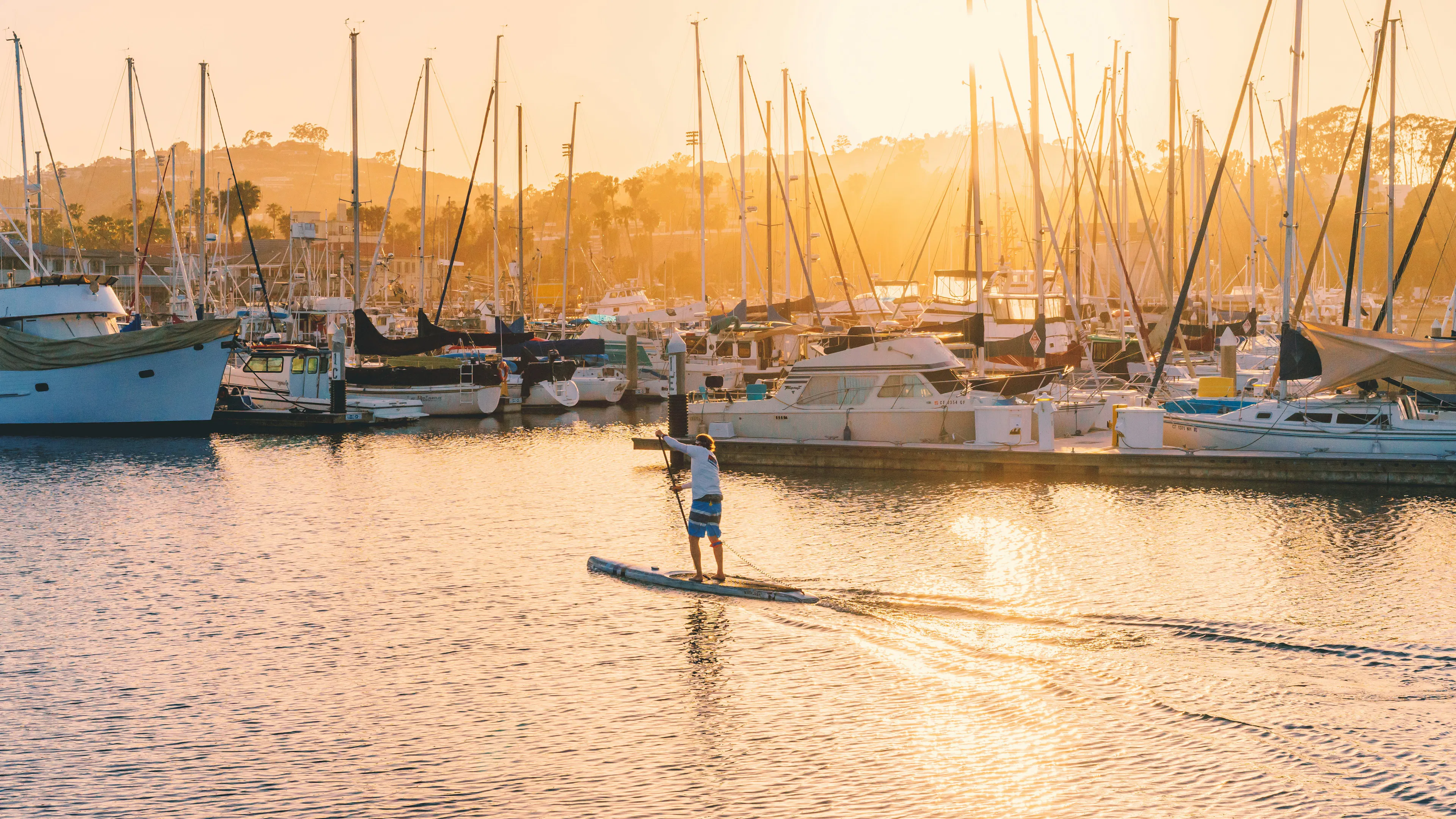 A stand up paddle boarder gliding past some boats in the sunset