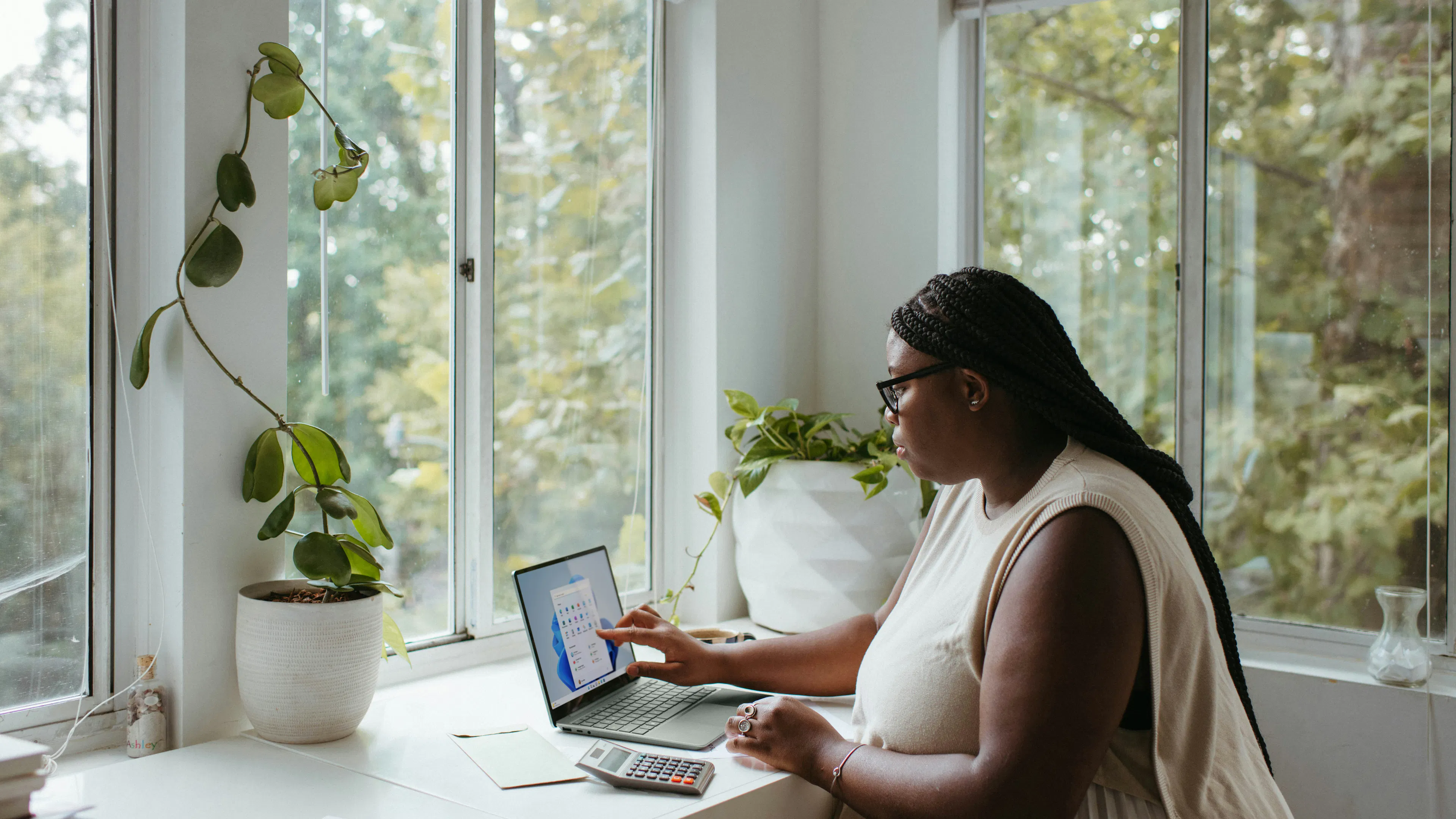 A woman sitting at a desk working on her laptop surrounded by green plants and bright windows