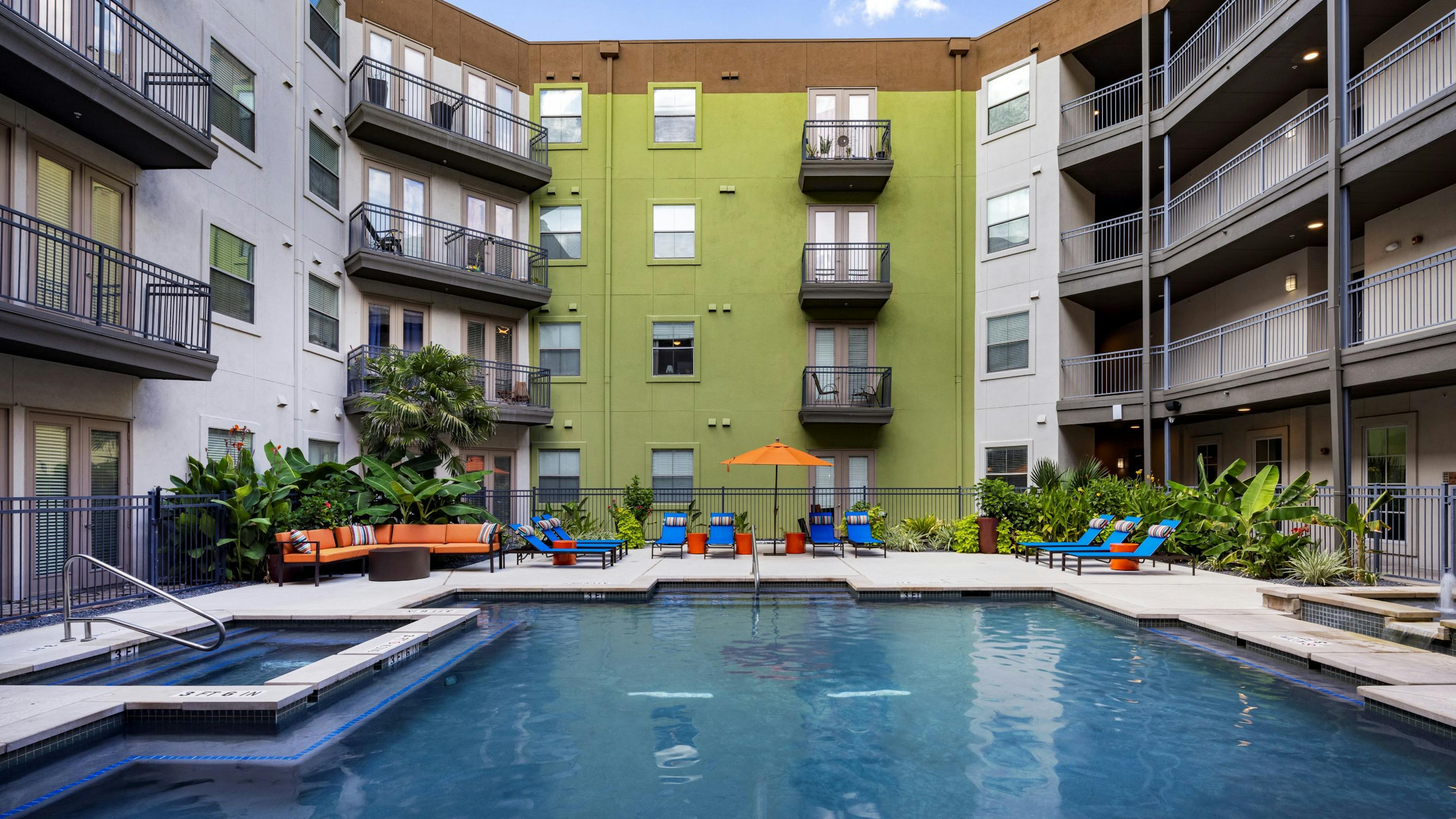 Daytime view of AMI 300 courtyard swimming pool with blue lounge chairs, an orange umbrella and couch and greenery