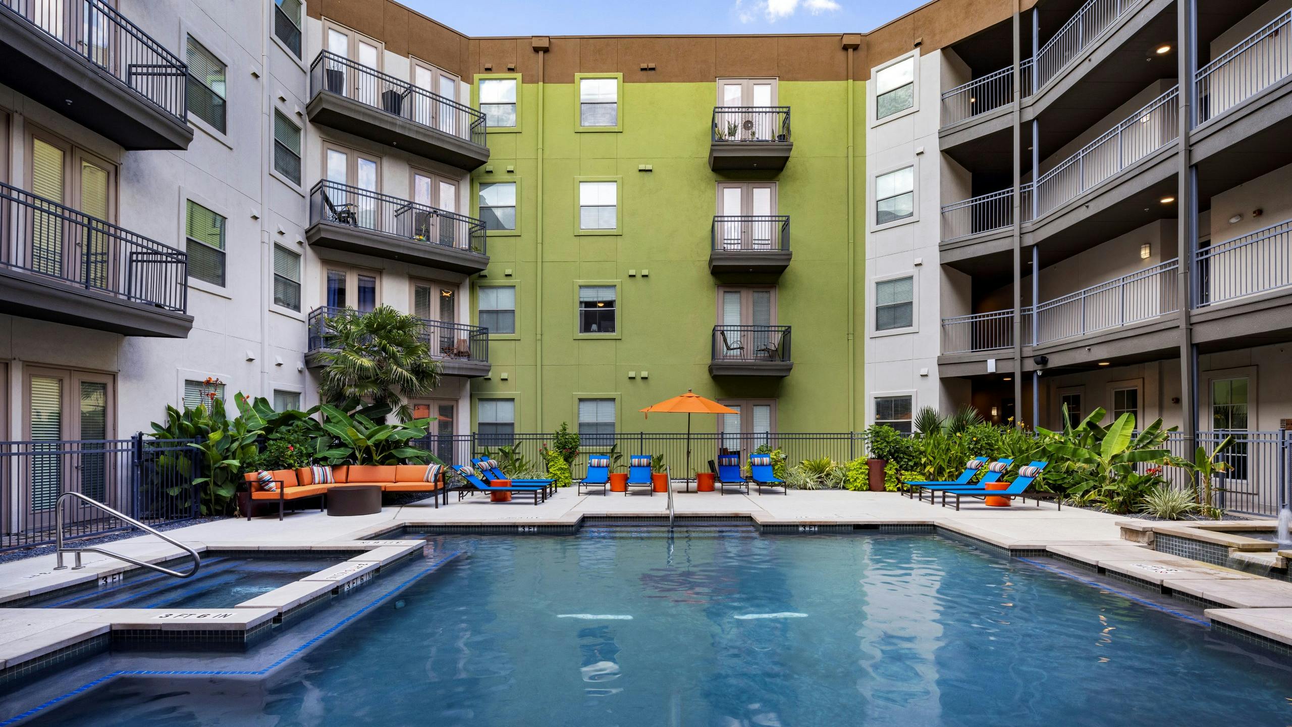 Daytime view of AMI 300 courtyard swimming pool with blue lounge chairs, an orange umbrella and couch and greenery