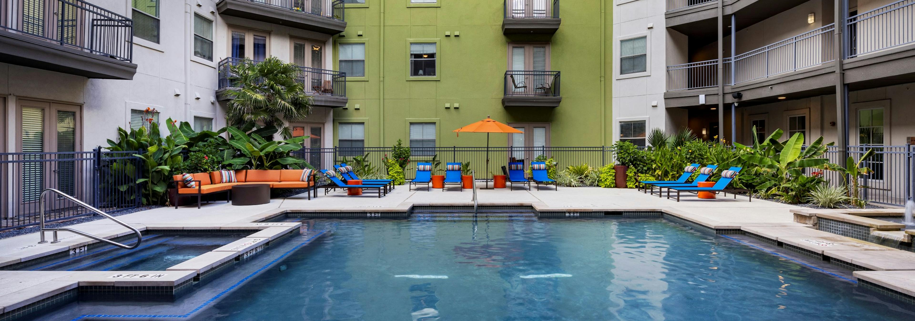 Daytime view of AMI 300 courtyard swimming pool with blue lounge chairs, an orange umbrella and couch and greenery