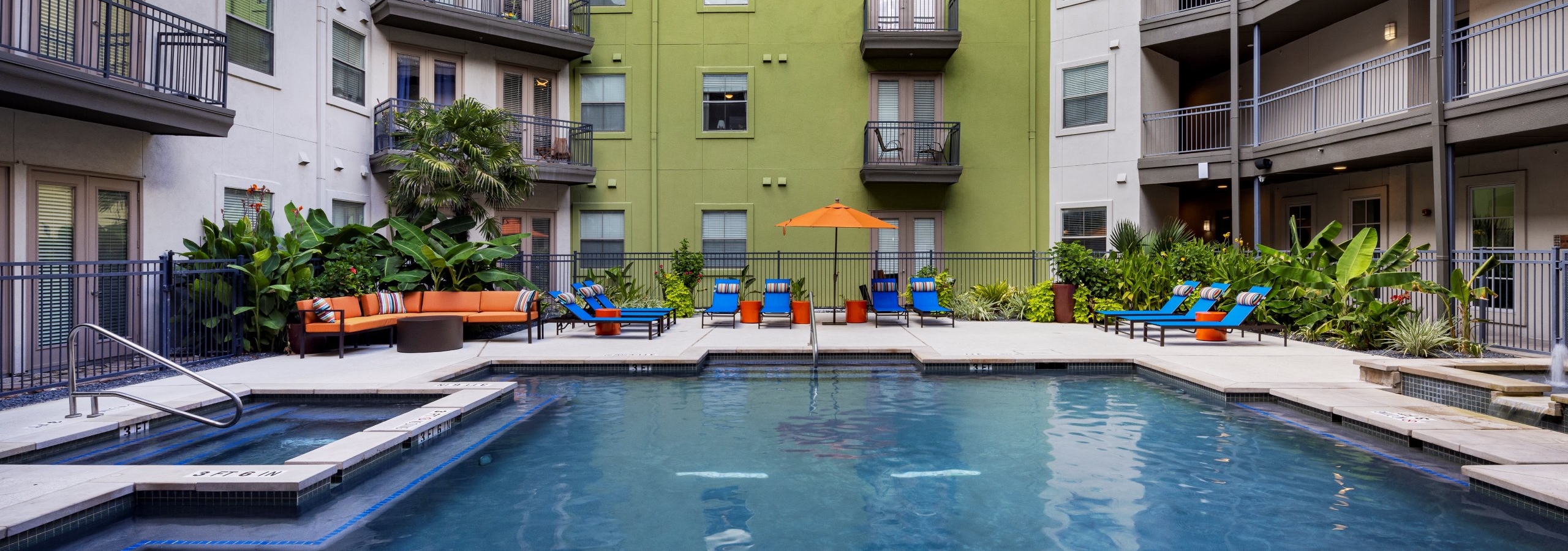 Daytime view of AMI 300 courtyard swimming pool with blue lounge chairs, an orange umbrella and couch and greenery