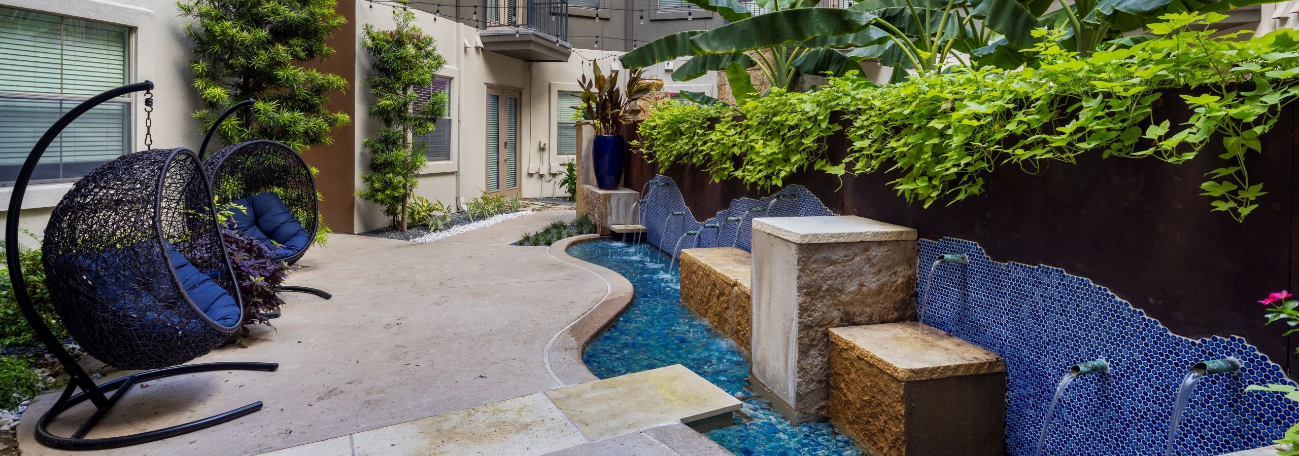 Daytime view of courtyard with a winding blue tile fountain and wicker hanging chairs with lush greenery at AMLI 300