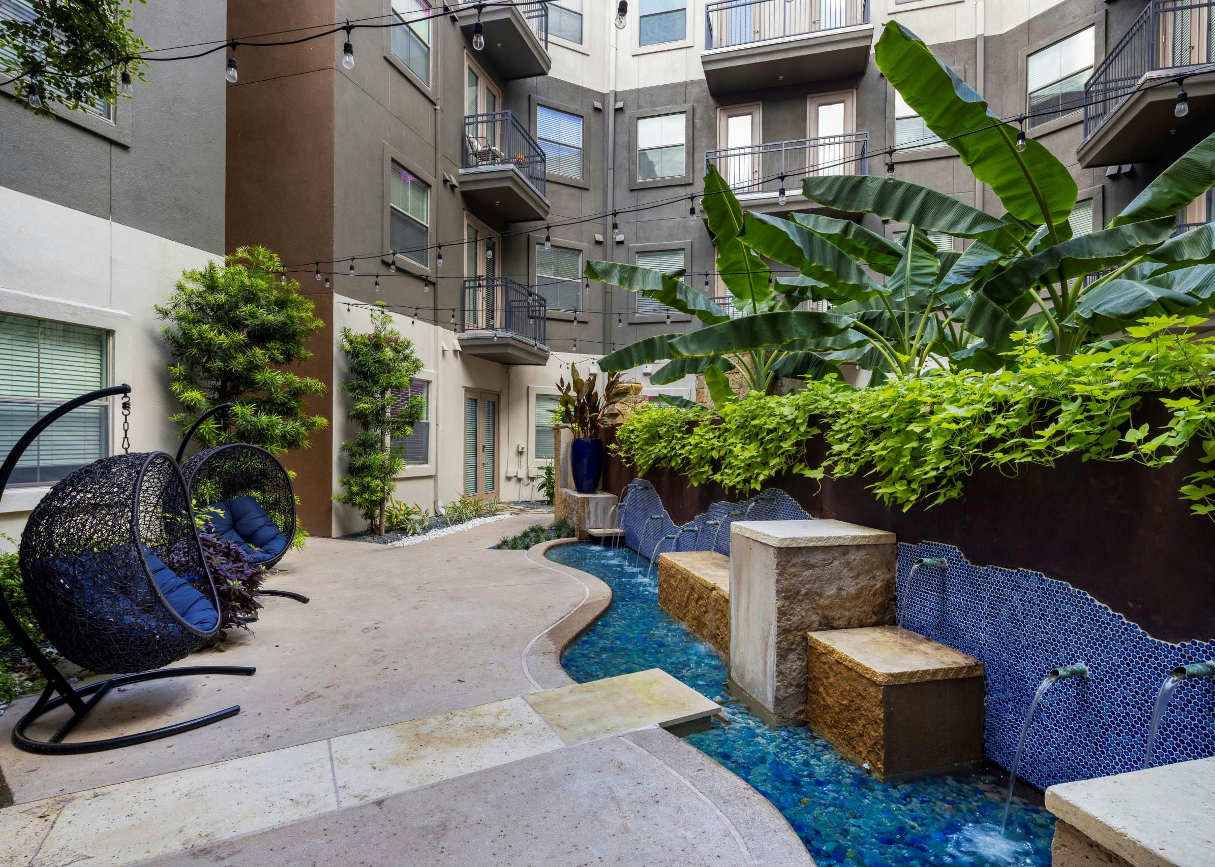 Daytime view of courtyard with a winding blue tile fountain and wicker hanging chairs with lush greenery at AMLI 300