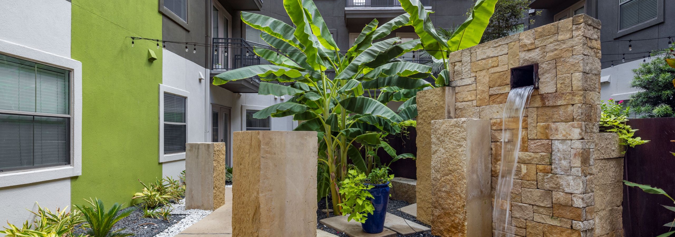An outdoor courtyard at AMLI 300 apartments featuring a sleek waterfall fountain with stacked beige stone design and lush banana plants