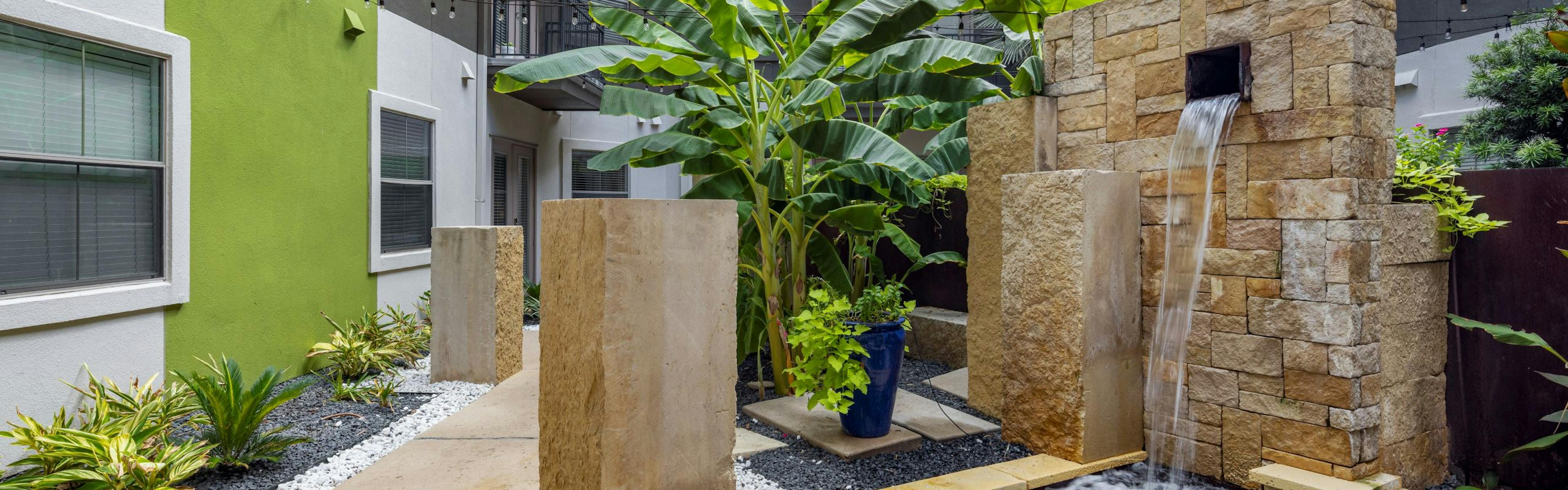An outdoor courtyard at AMLI 300 apartments featuring a sleek waterfall fountain with stacked beige stone design and lush banana plants