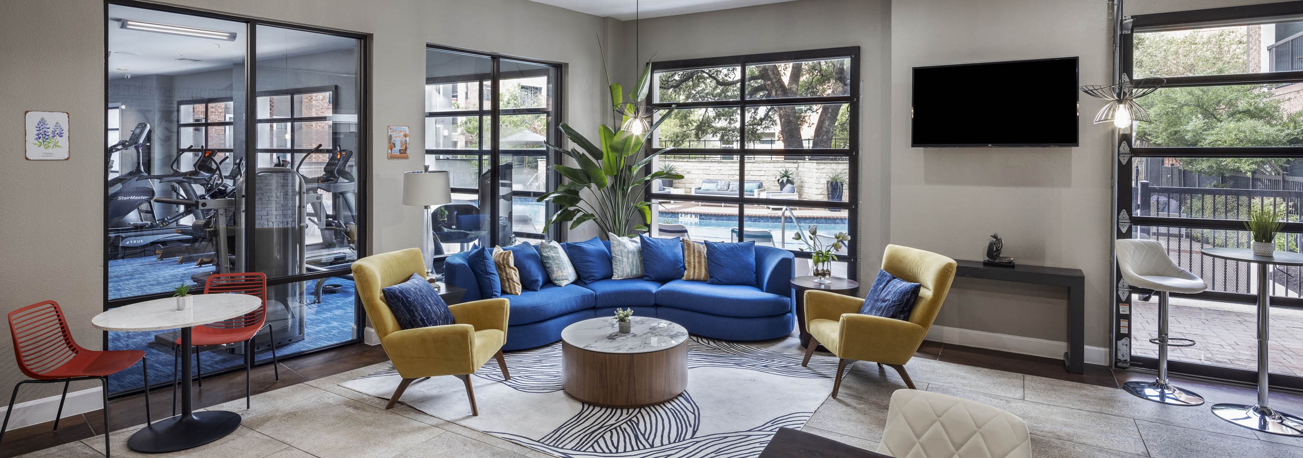 AMLI Eastside resident lounge with a blue sectional sofa and yellow armchairs and the gym and pool are visible through windows
