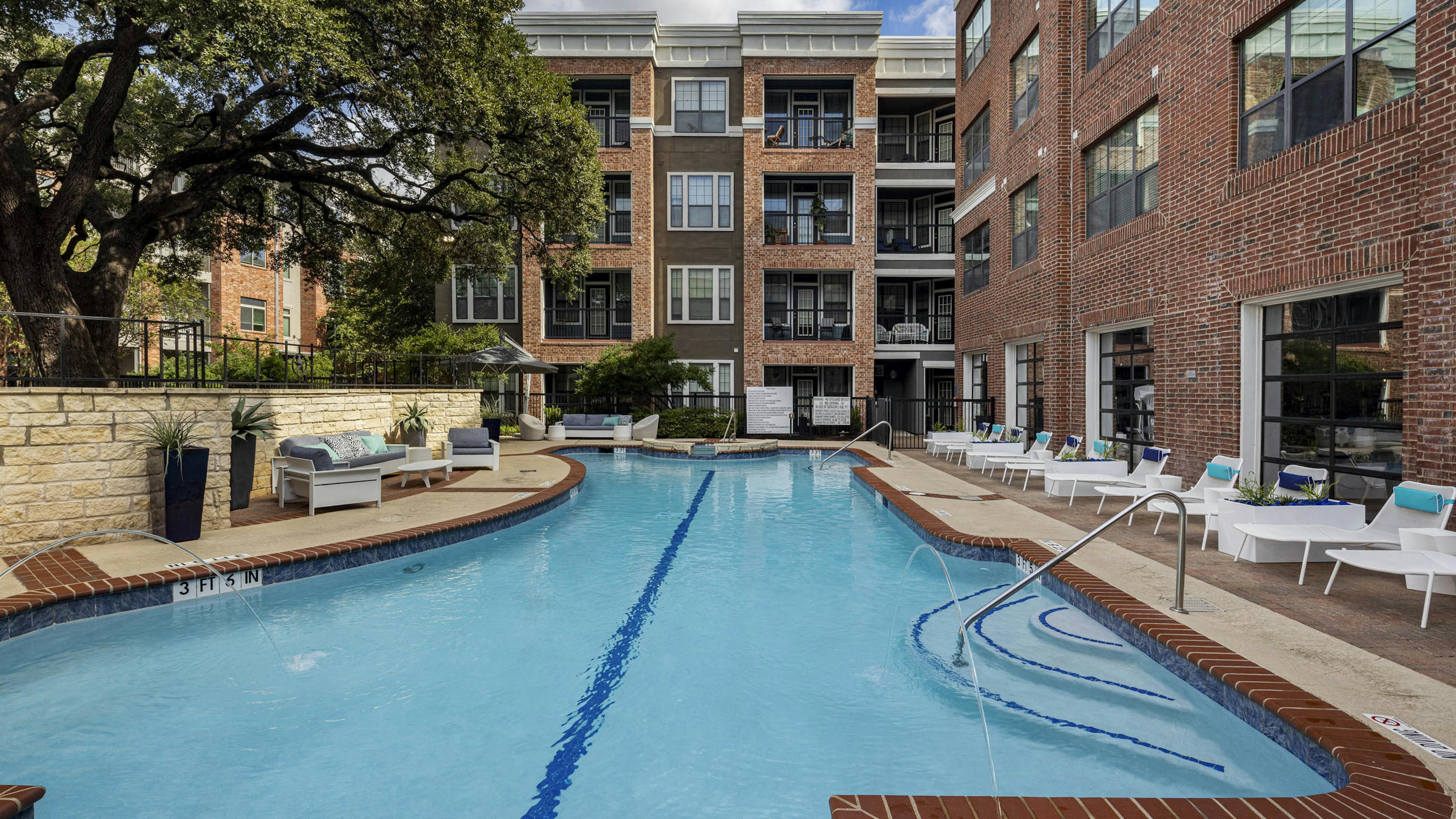 Day view of swimming pool at AMLI Eastside apartments with white lounge chairs, cushioned seating, brick buildings and a large tree