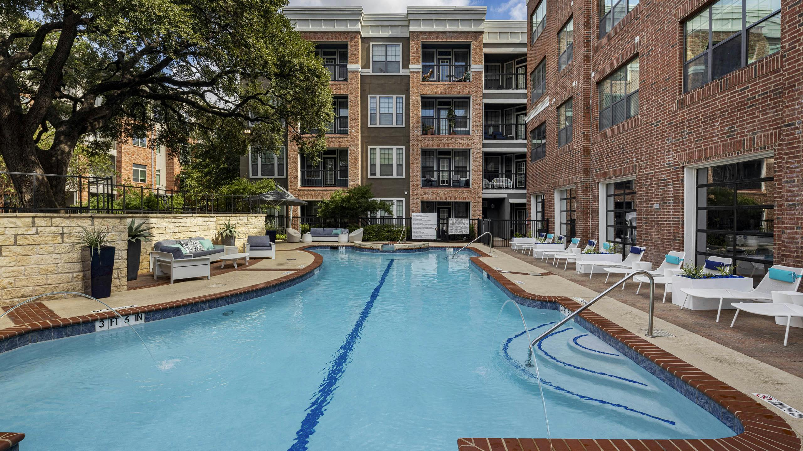 Day view of swimming pool at AMLI Eastside apartments with white lounge chairs, cushioned seating, brick buildings and a large tree
