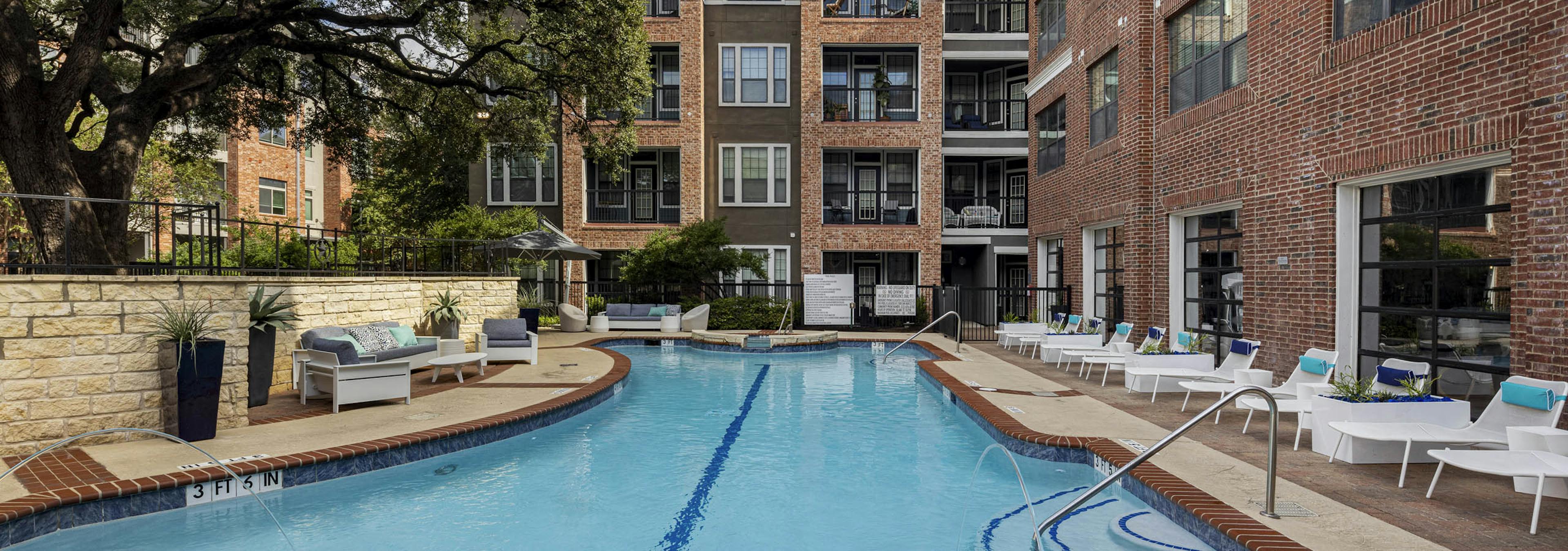 Day view of swimming pool at AMLI Eastside apartments with white lounge chairs, cushioned seating, brick buildings and a large tree