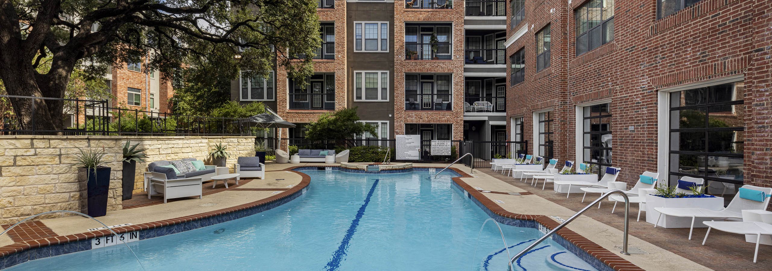 Day view of swimming pool at AMLI Eastside apartments with white lounge chairs, cushioned seating, brick buildings and a large tree