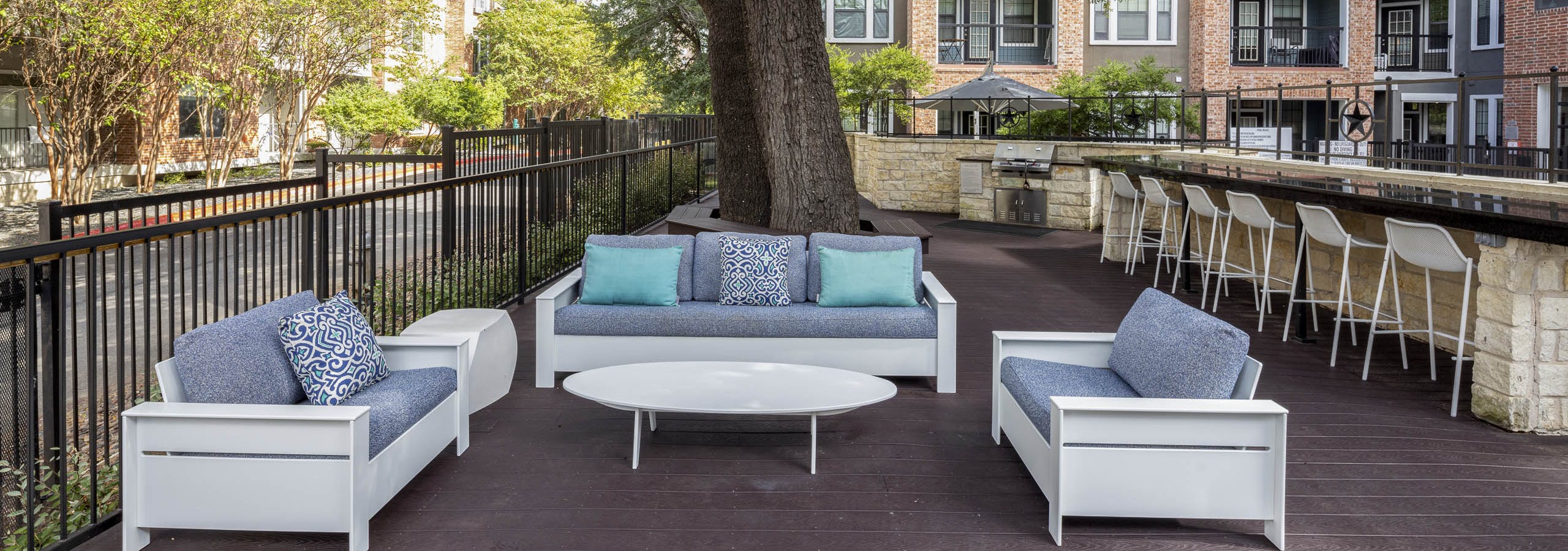 Poolside grill deck at AMLI Eastside apartments with cushioned seating and a long counter with 7 white bar stools shaded by a large tree