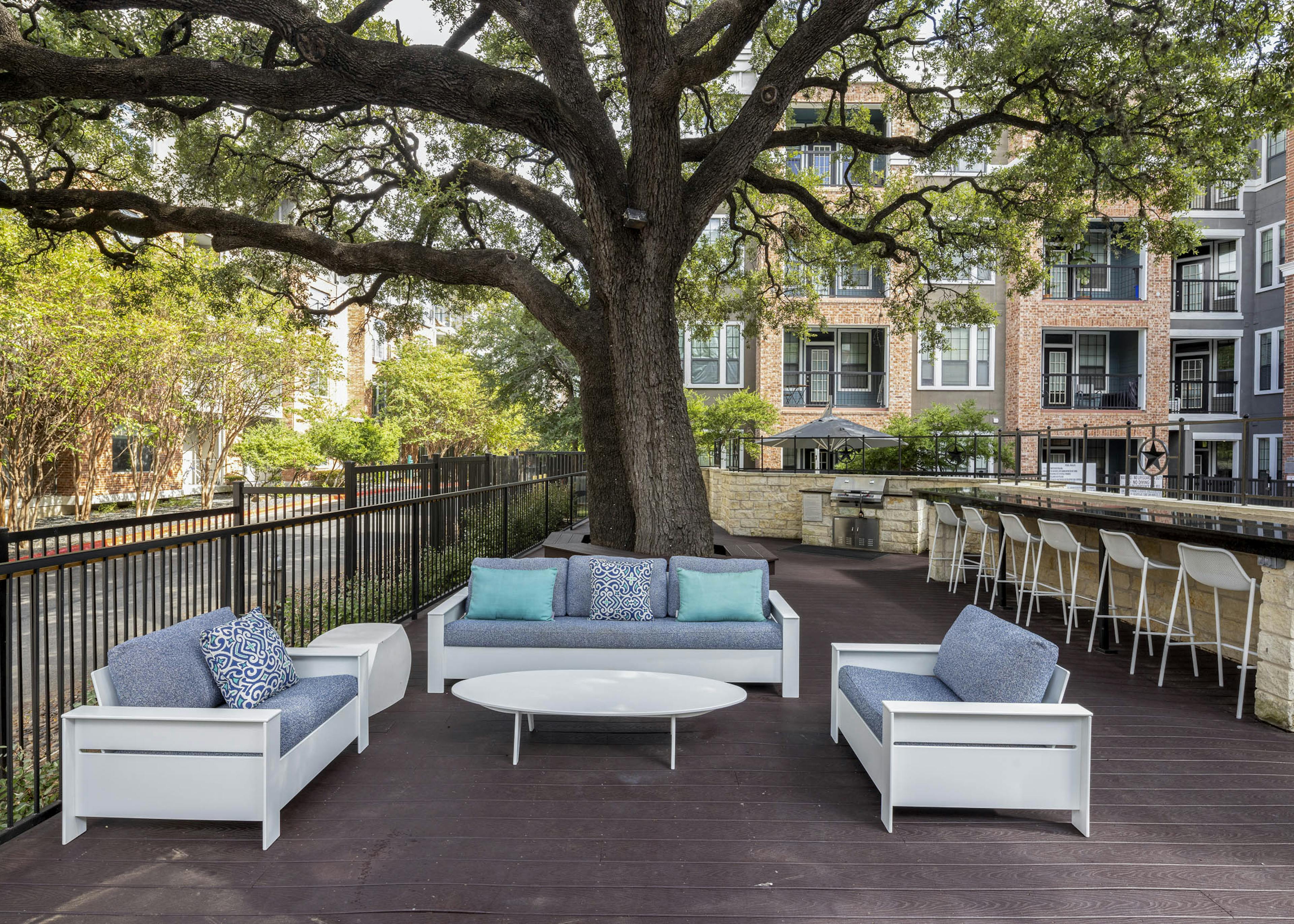 Poolside grill deck at AMLI Eastside apartments with cushioned seating and a long counter with 7 white bar stools shaded by a large tree
