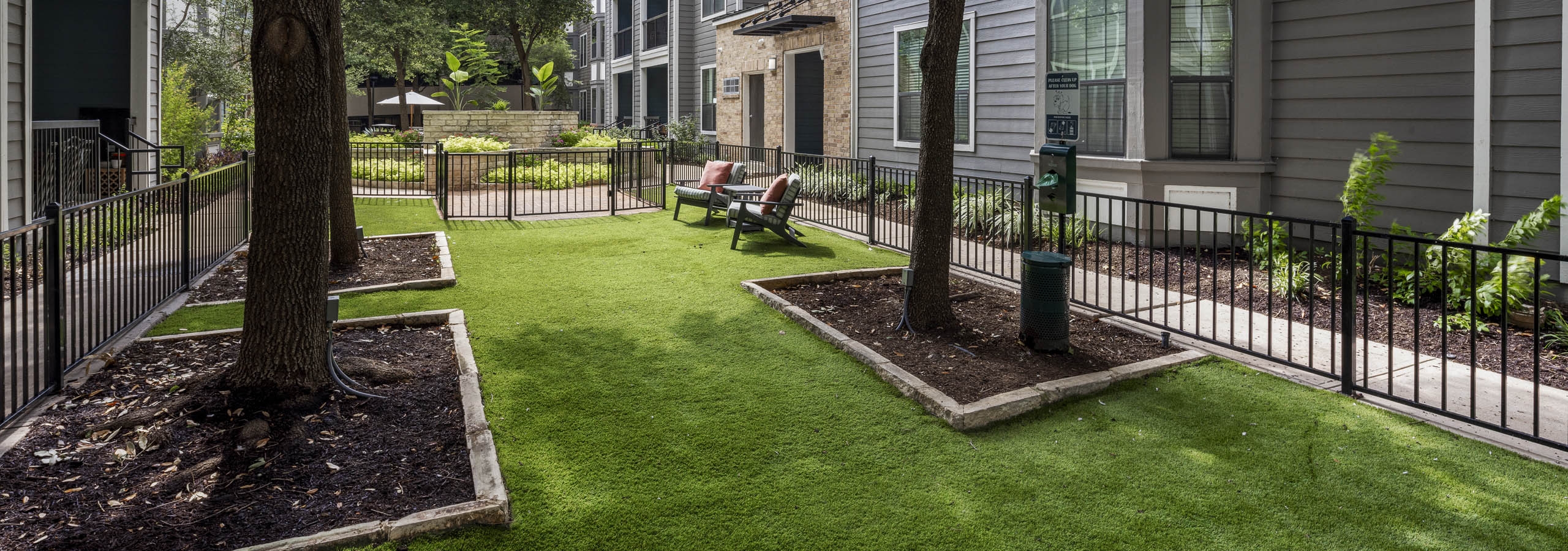 Daytime view of a fenced grassy courtyard featuring two lounge chairs, mature trees, and a dog waste station at AMLI Eastside apartments