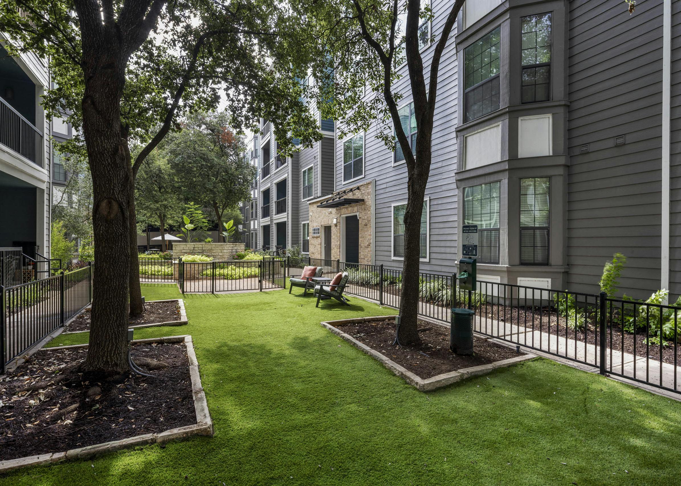 Daytime view of a fenced grassy courtyard featuring two lounge chairs, mature trees, and a dog waste station at AMLI Eastside apartments
