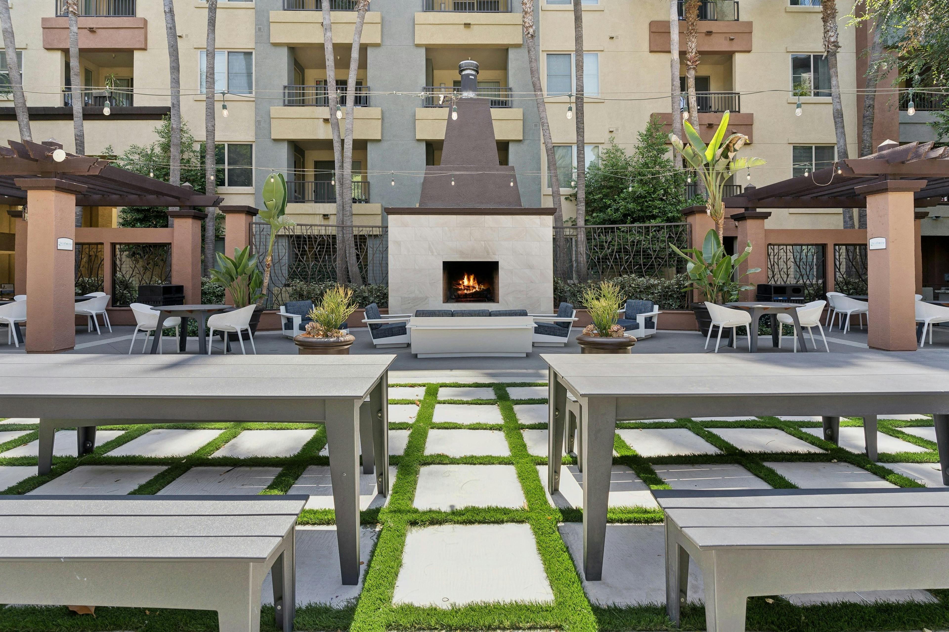 Outdoor area with long gray tables and benches on a grass-tiled pathway that leads to a lit fireplace at AMLI Warner Center apartments