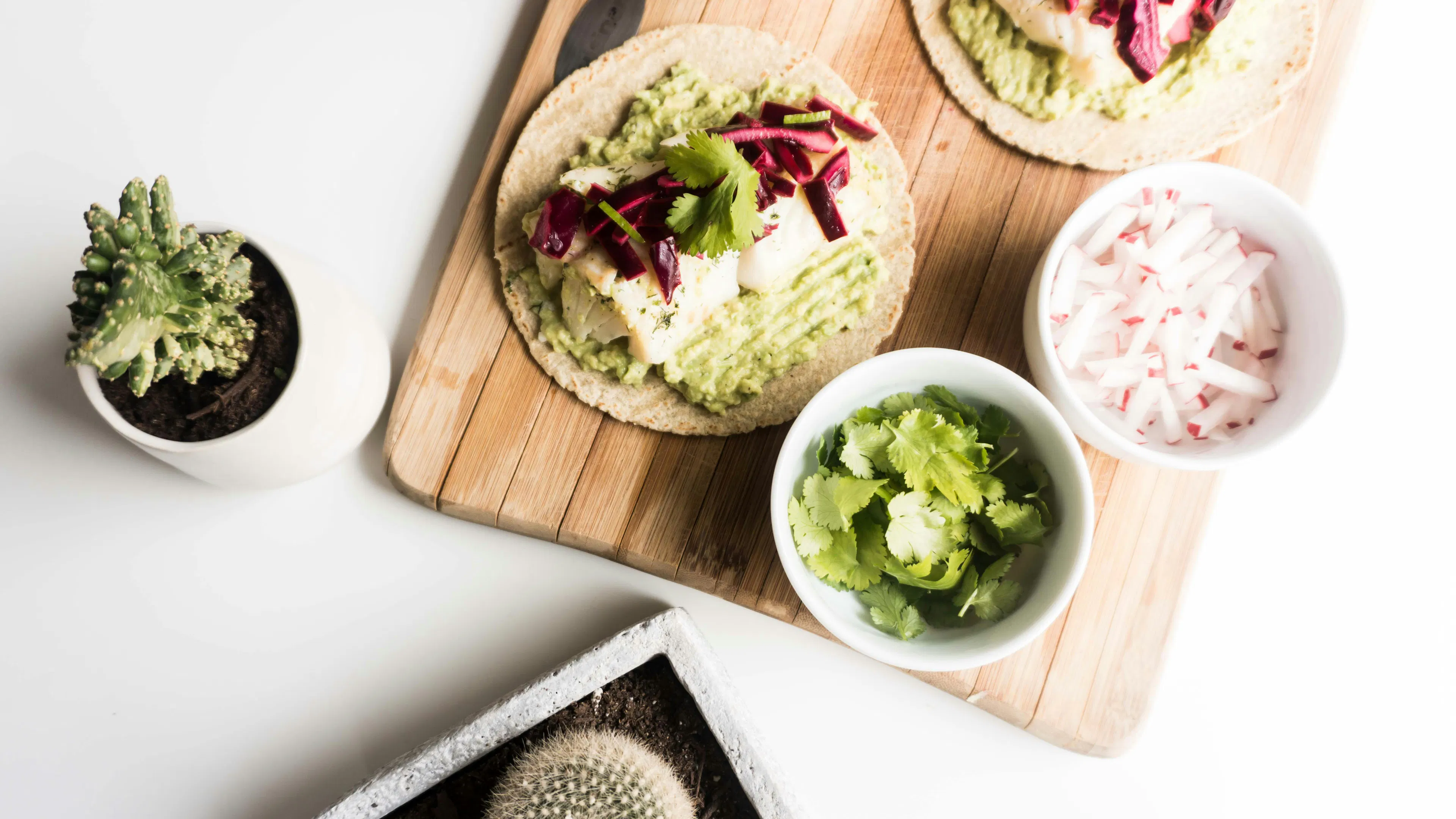 A fish taco on a wooden board next to bowls of avocado and onion