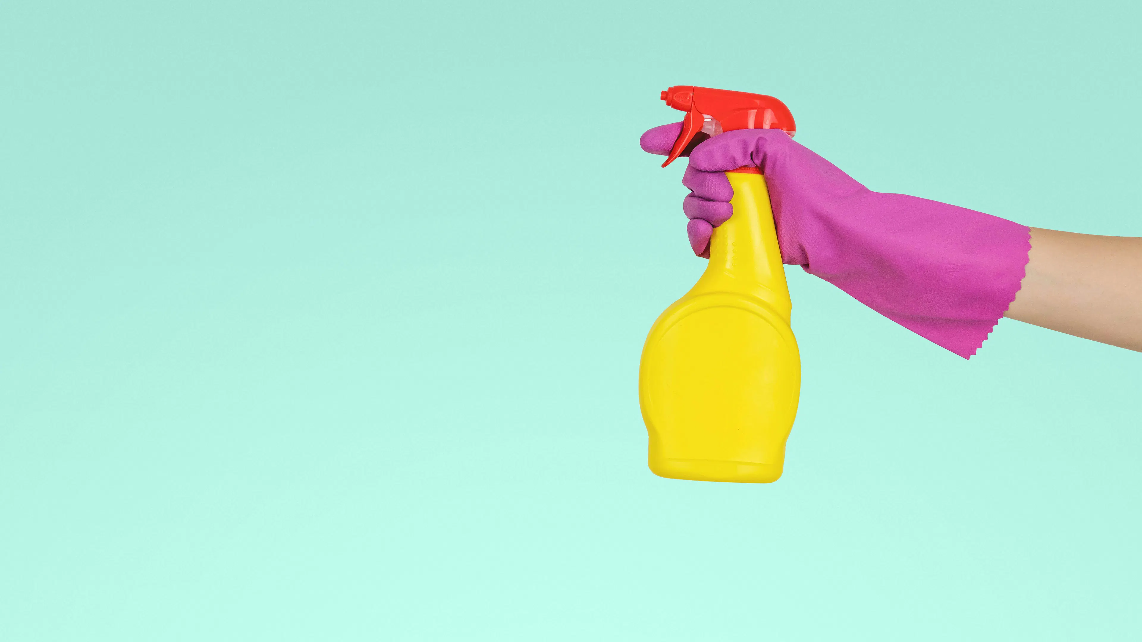 A pink gloved hand holding a yellow spray bottle over a light green background