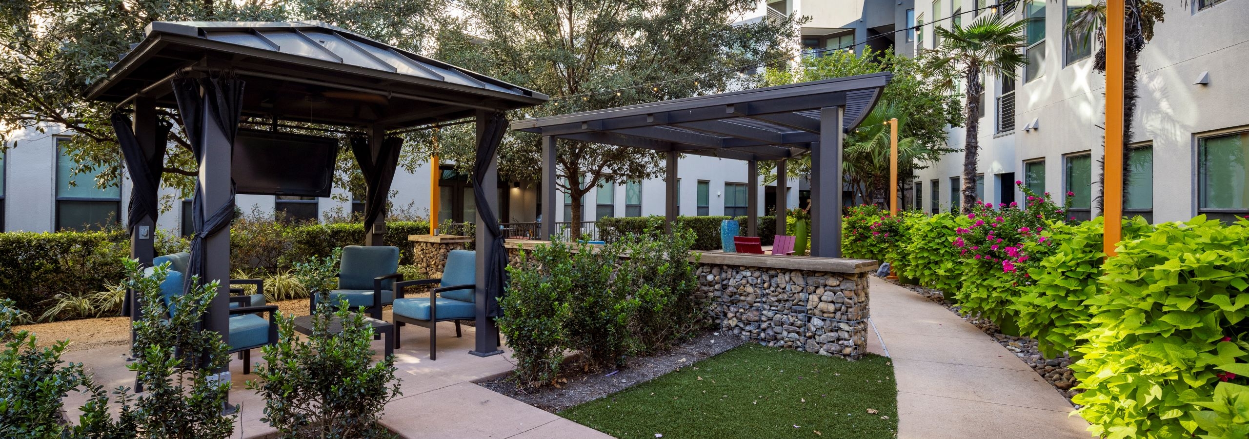 Day view of AMLI on Aldrich courtyard boasts lush greenery, two inviting pergolas with seating areas and a TV hanging beneath one.