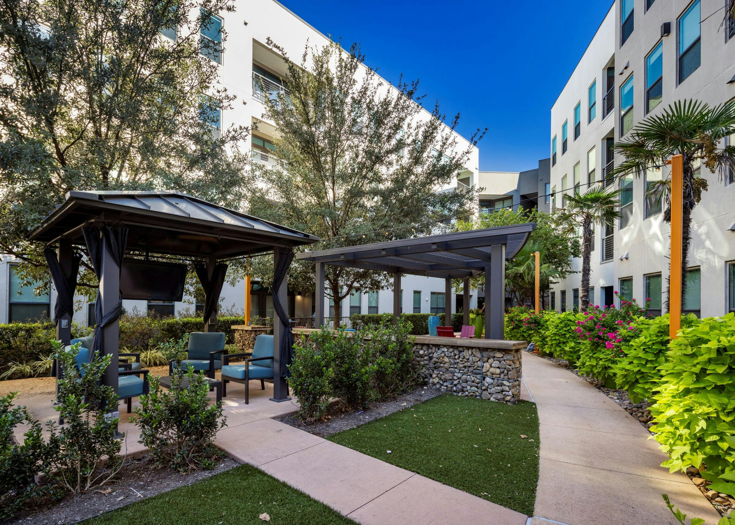 Day view of AMLI on Aldrich courtyard boasts lush greenery, two inviting pergolas with seating areas and a TV hanging beneath one.