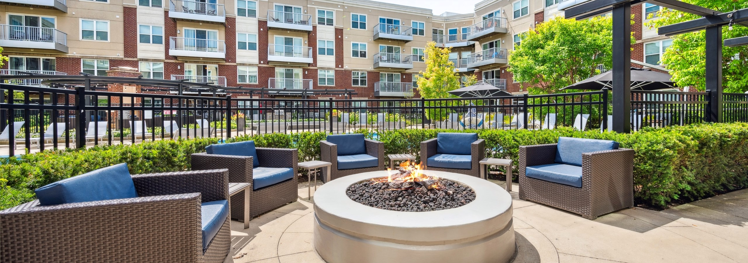 Exterior view of fire pit lounge area surrounded by wicker chairs in front of pool gate with apartments in background at AMLI Deerfield.