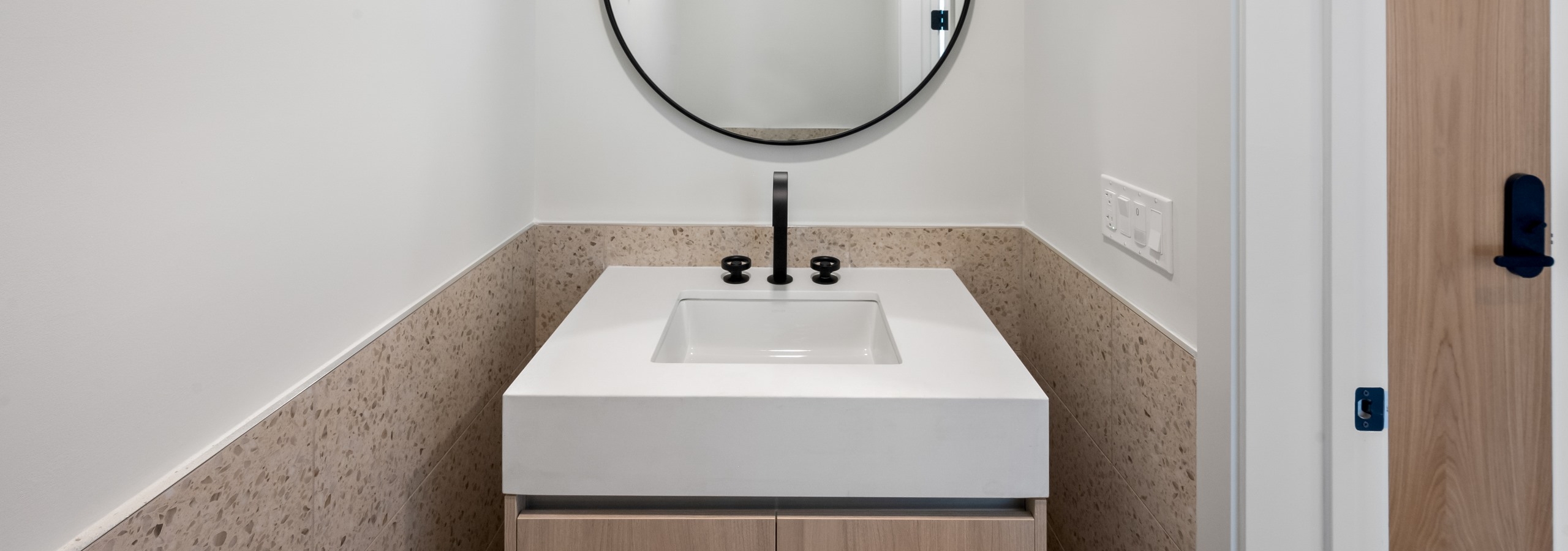 AMLI Wynwood powder room with modern, light wood vanity, square white top, black faucet and round mirror above half tiled wall