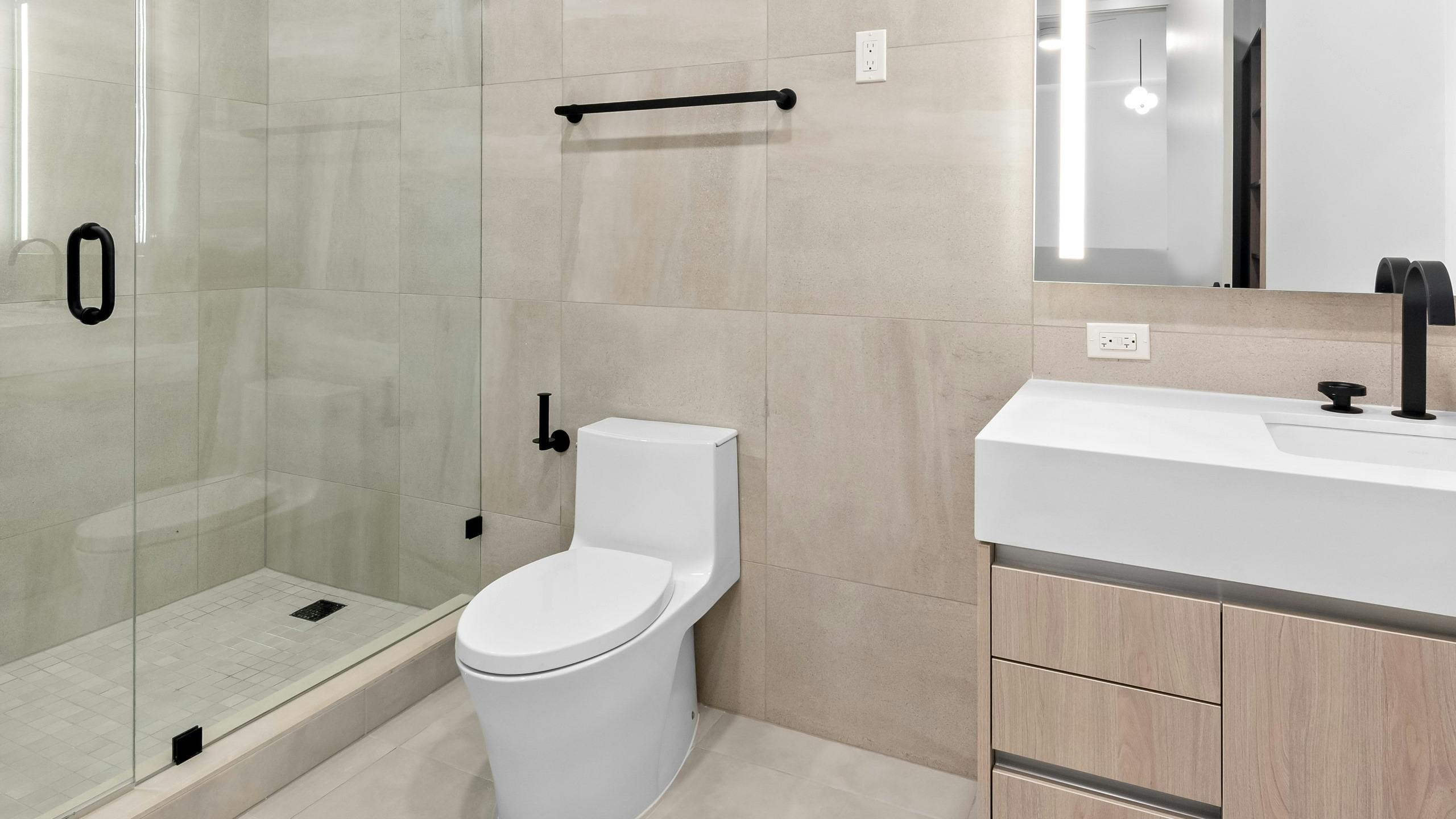 AMLI Wynwood modern bathroom with glass shower, white toilet, beige tile walls and floor and light wood vanity with thick white stone top.