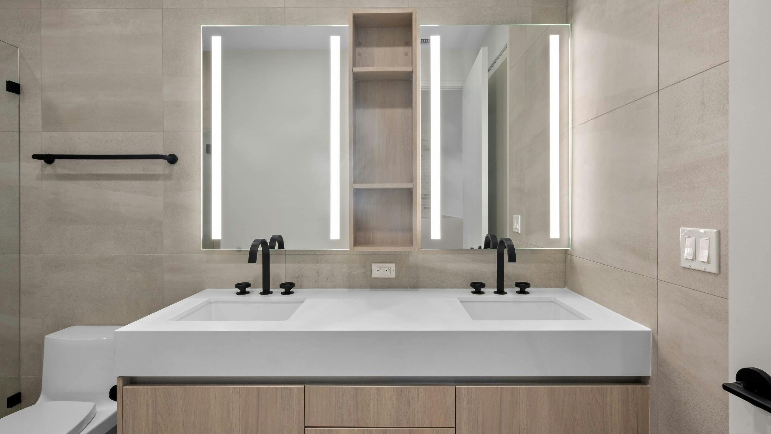 AMLI Wynwood bathroom with beige tile walls, light wood double vanity with thick white stone top and lit mirrors above.