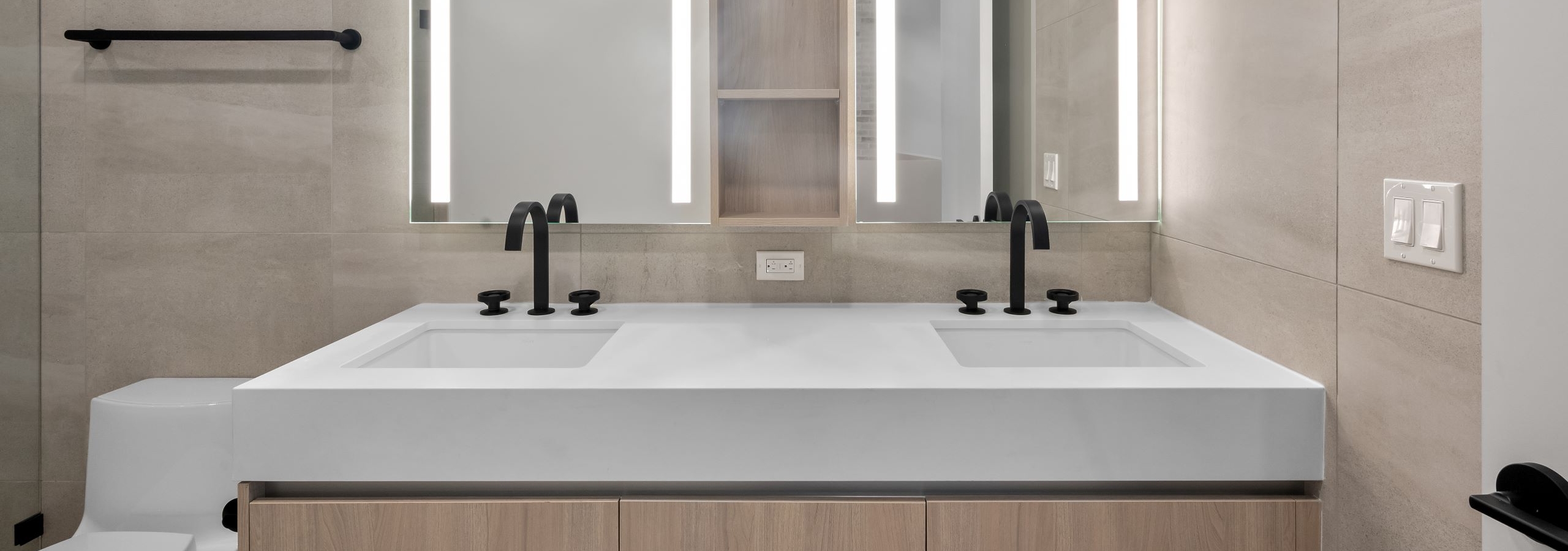 AMLI Wynwood bathroom with beige tile walls, light wood double vanity with thick white stone top and lit mirrors above.