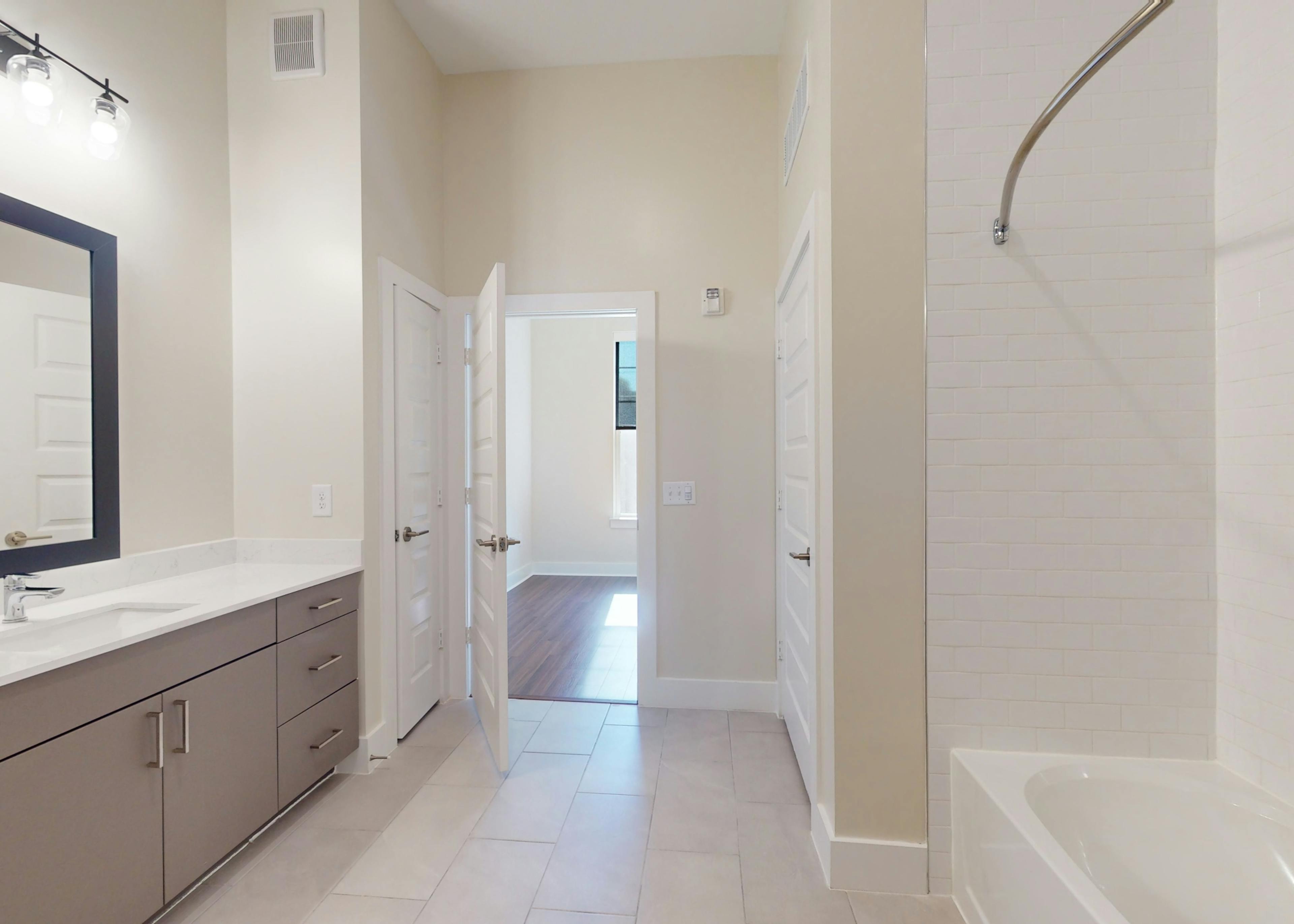AMLI Brookhaven bathroom with a soaking tub, modern gray vanity with mirror above, and tile floors looking onto a bedroom.