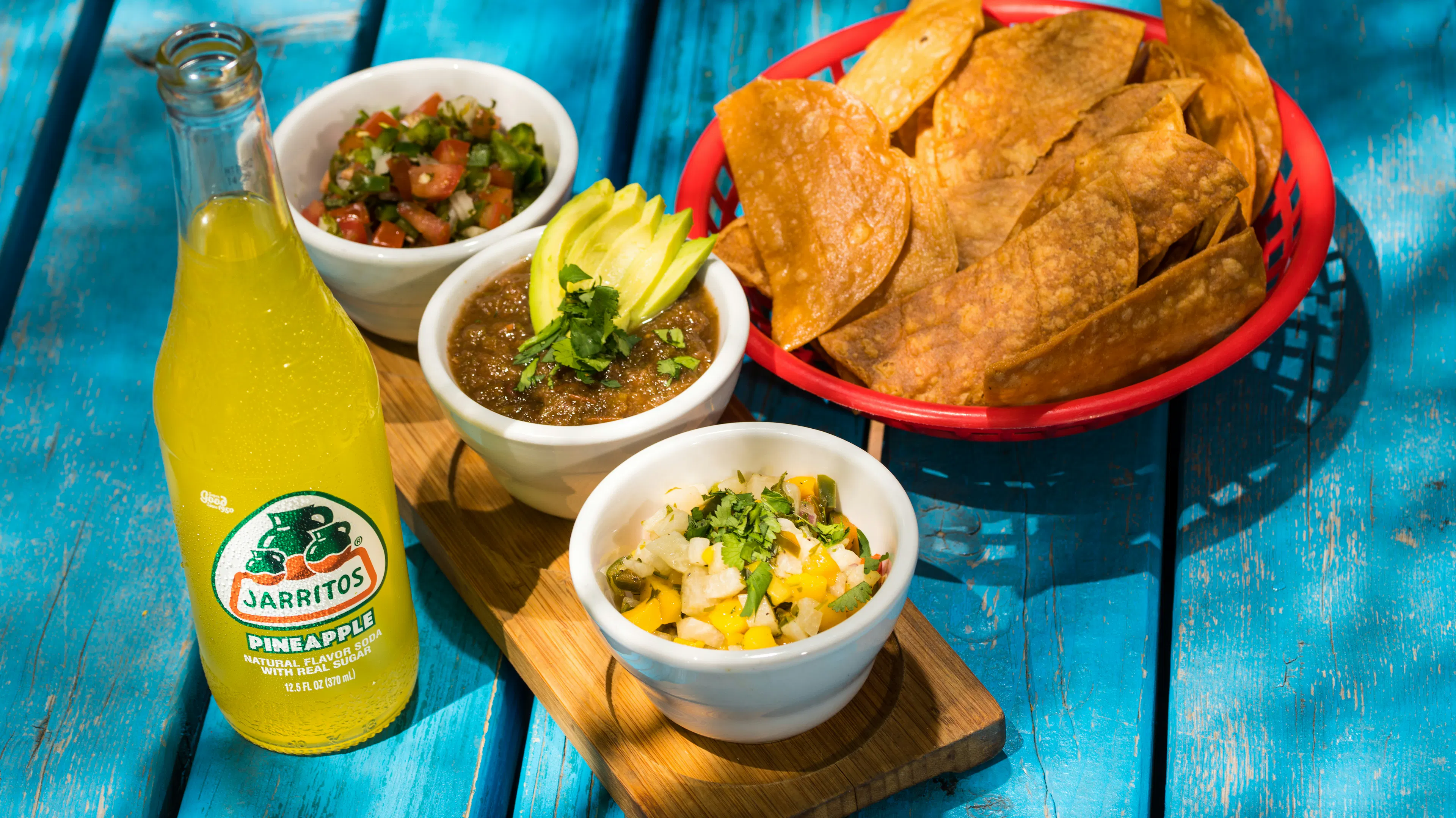 Three small bowls of different salsas beside a bowl of tortilla chips and a glass of soda
