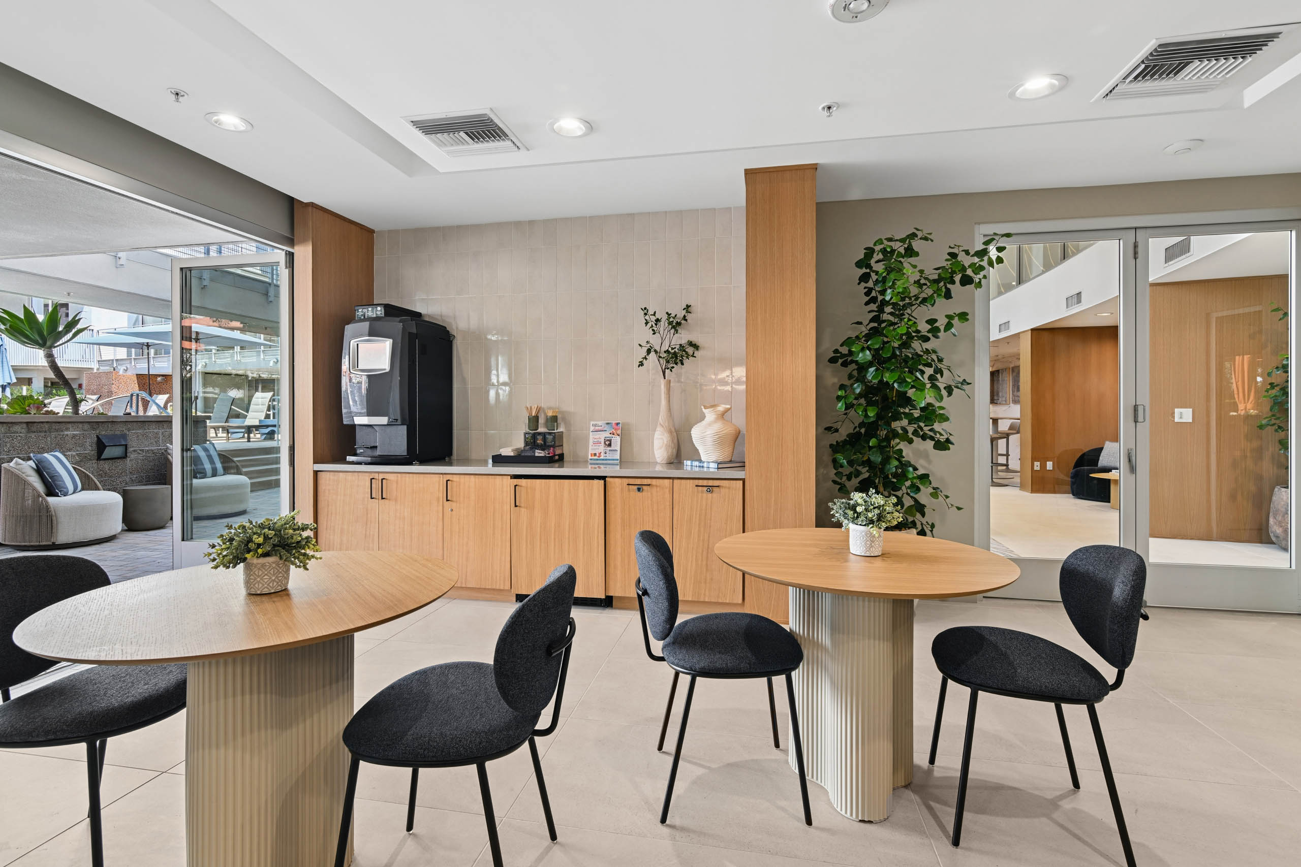 Social Hub at AMLI Lex on Orange with a coffee station, wood cabinetry, a tiled backsplash, round wood tables with dark chairs, and open doors leading to an outdoor lounge area.
