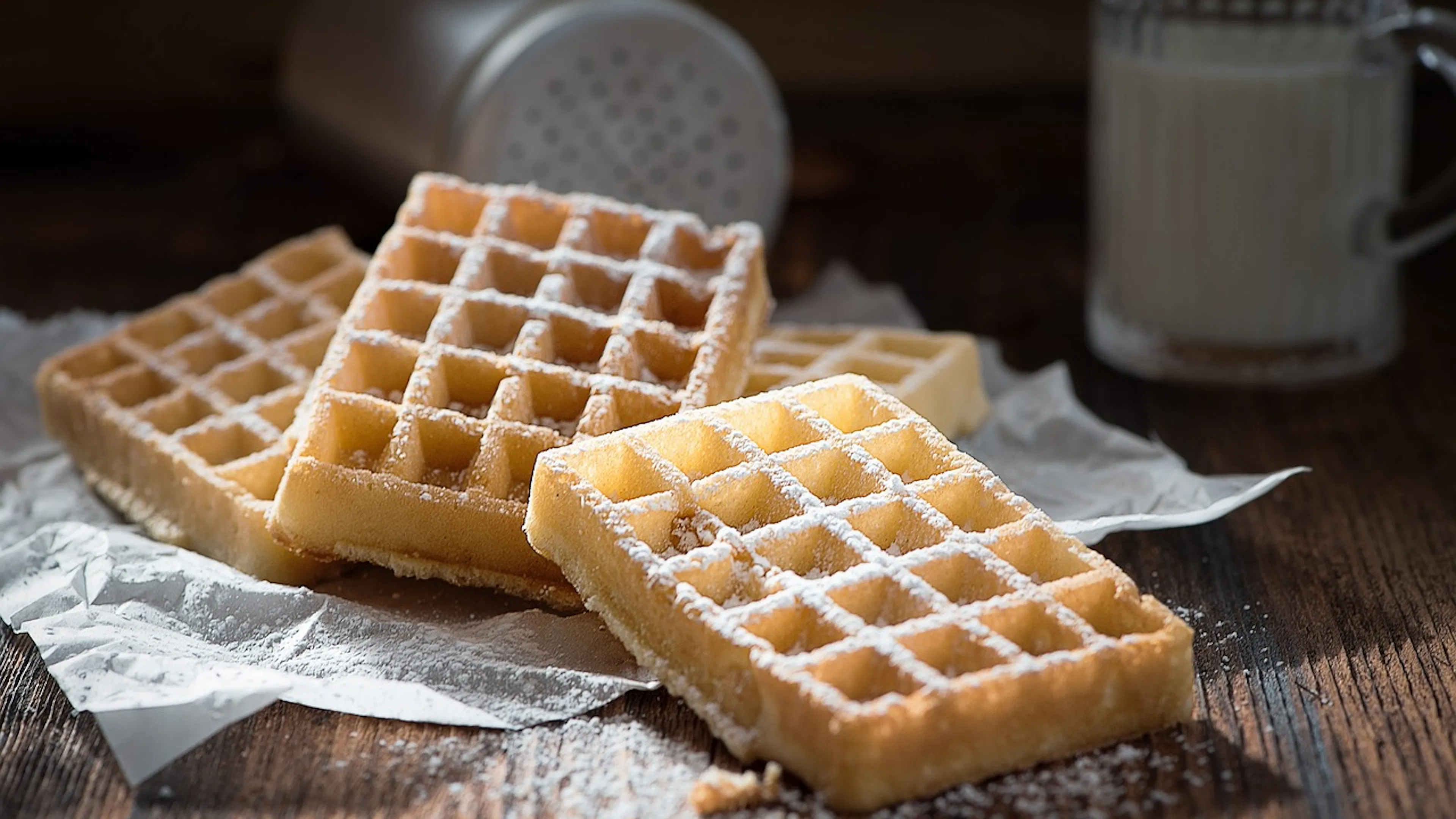 A stack of light rectangular waffles lightly dusted in powdered sugar sitting on top a dark wooden farmhouse style table