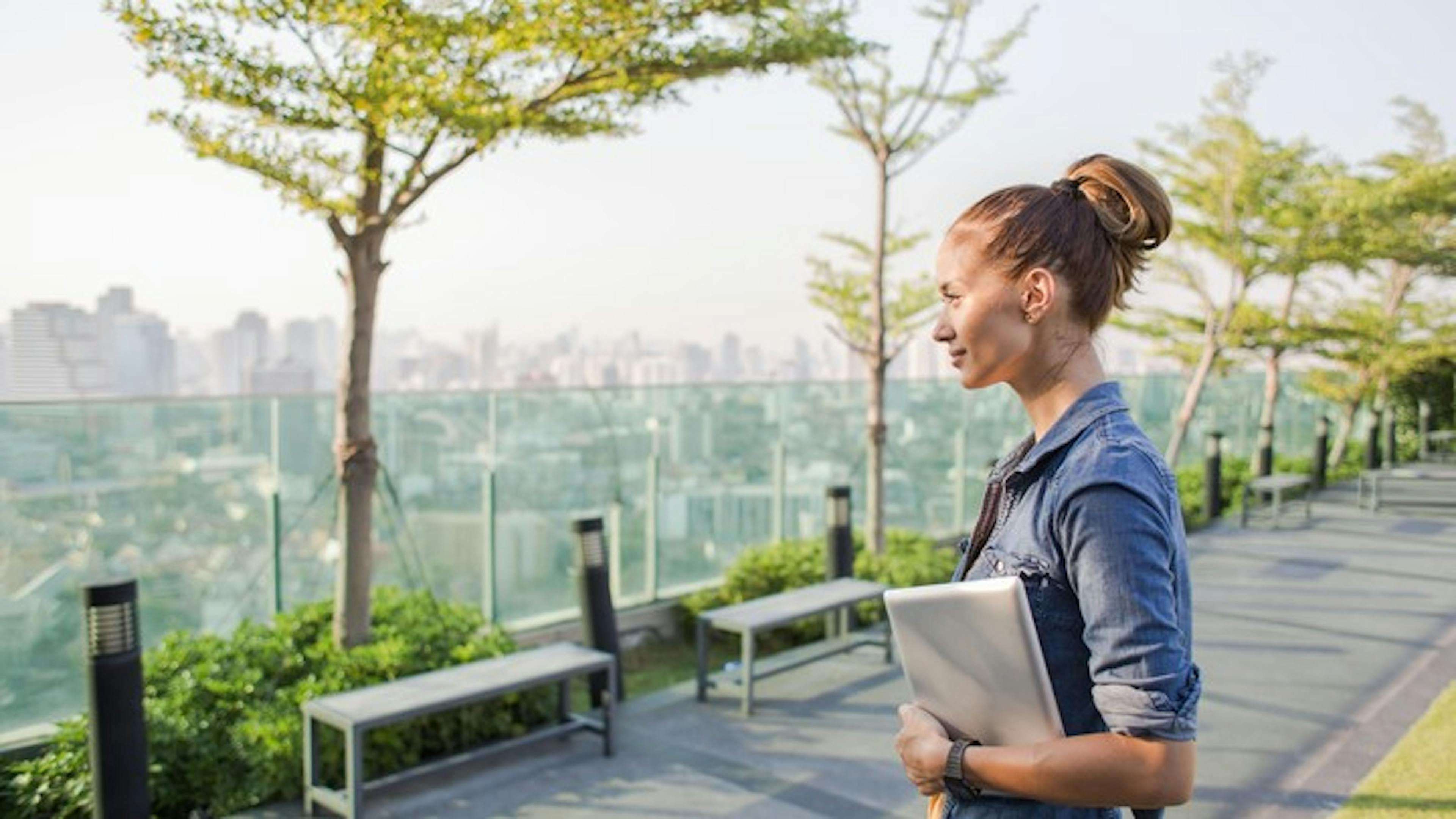 Woman on a skydeck holding her laptop and enjoying a bright daytime city view of buildings and green trees with a blue sky